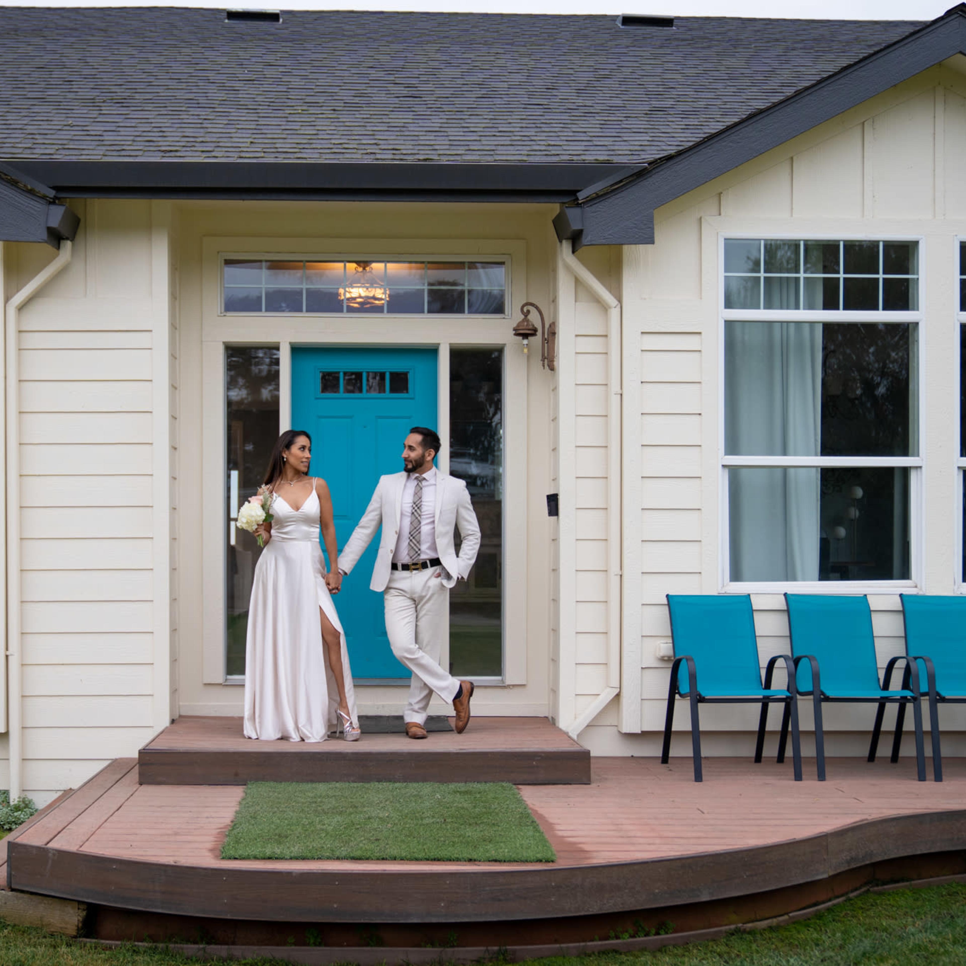 A couple dressed in formal attire is walking hand in hand on the porch of a house with teal accents and a row of blue chairs.