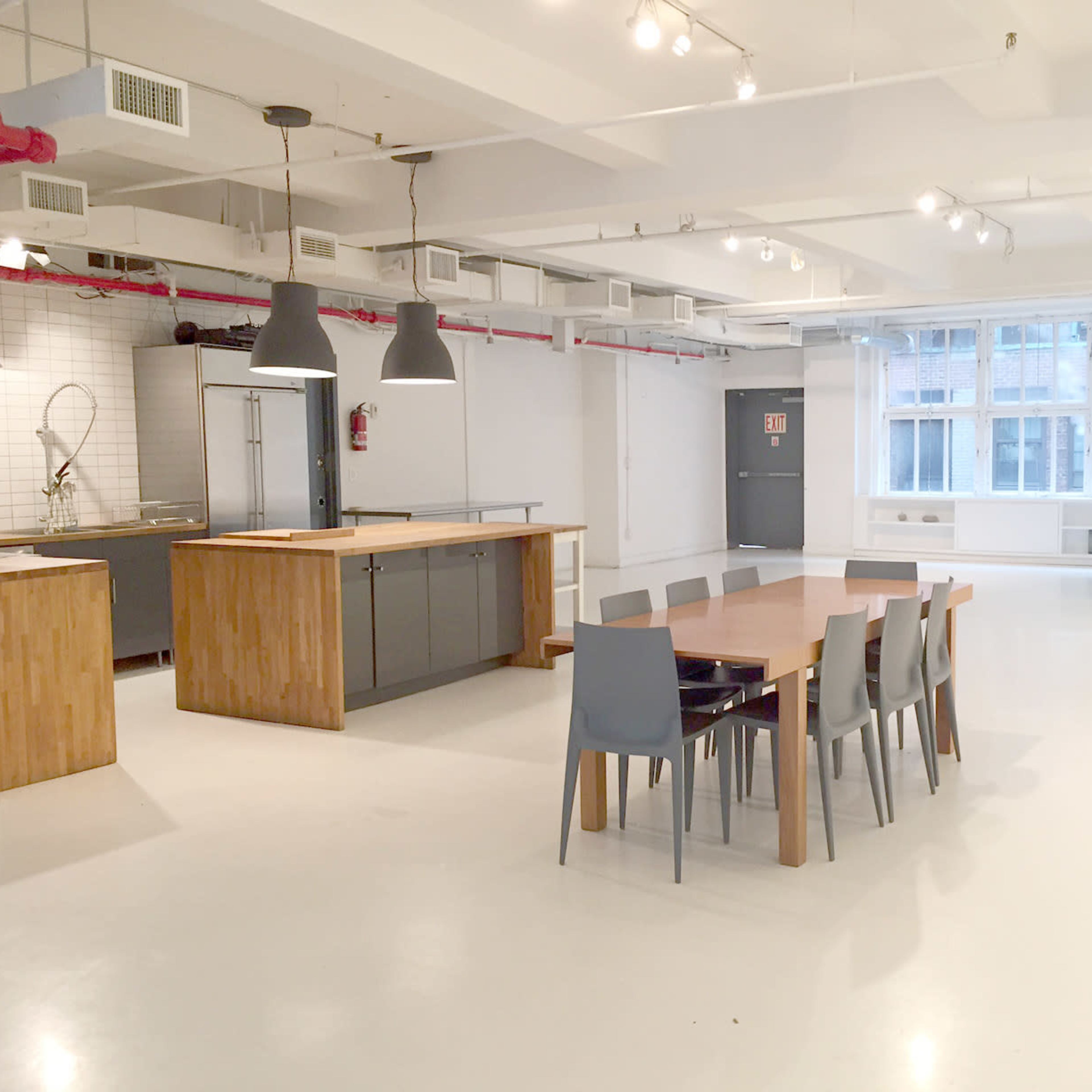 A modern kitchen and dining area featuring a long wooden table surrounded by six chairs, with large windows allowing natural light to fill the space.