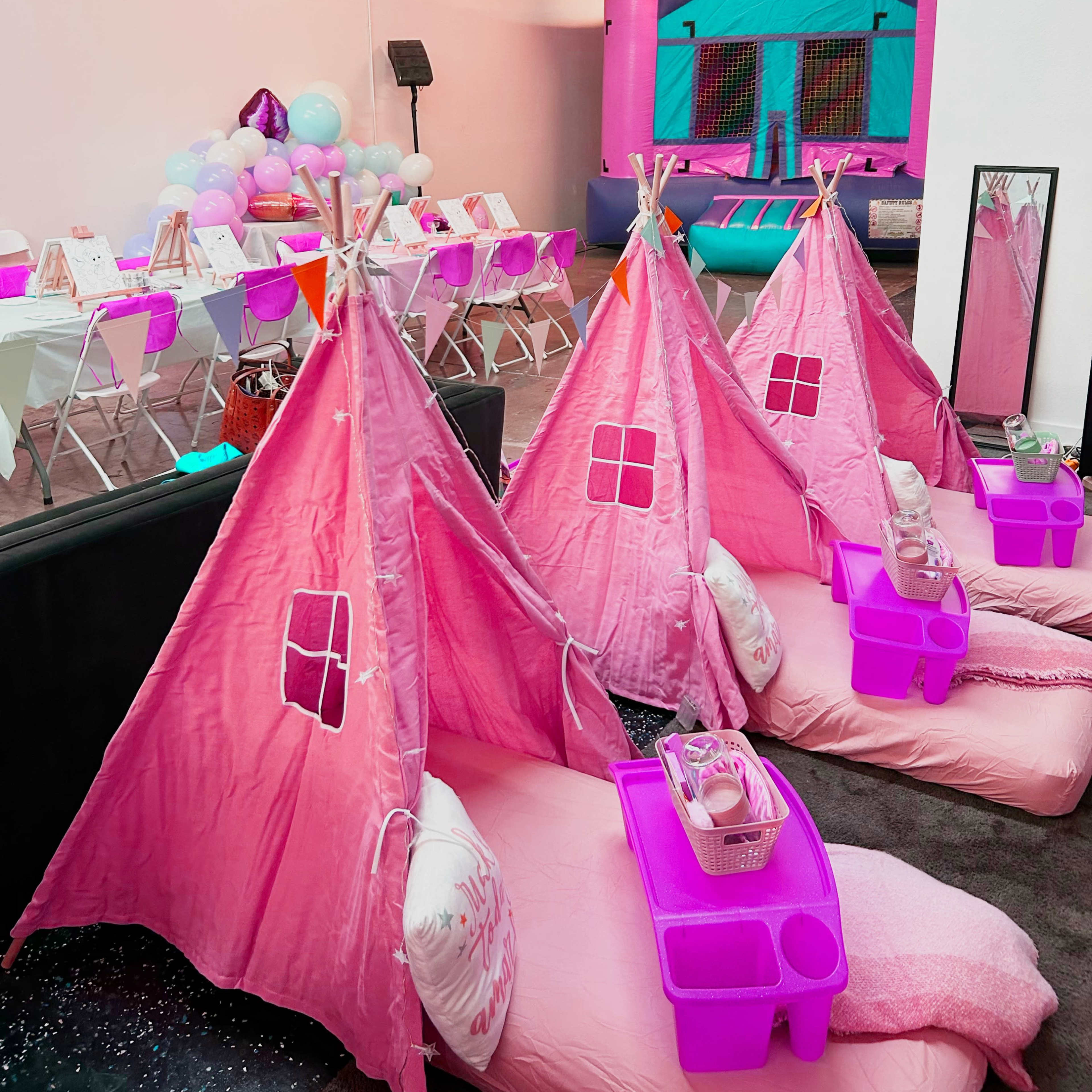 A children's party setup featuring pink teepees with mattresses and trays, complemented by a colorful bounce house in the background.
