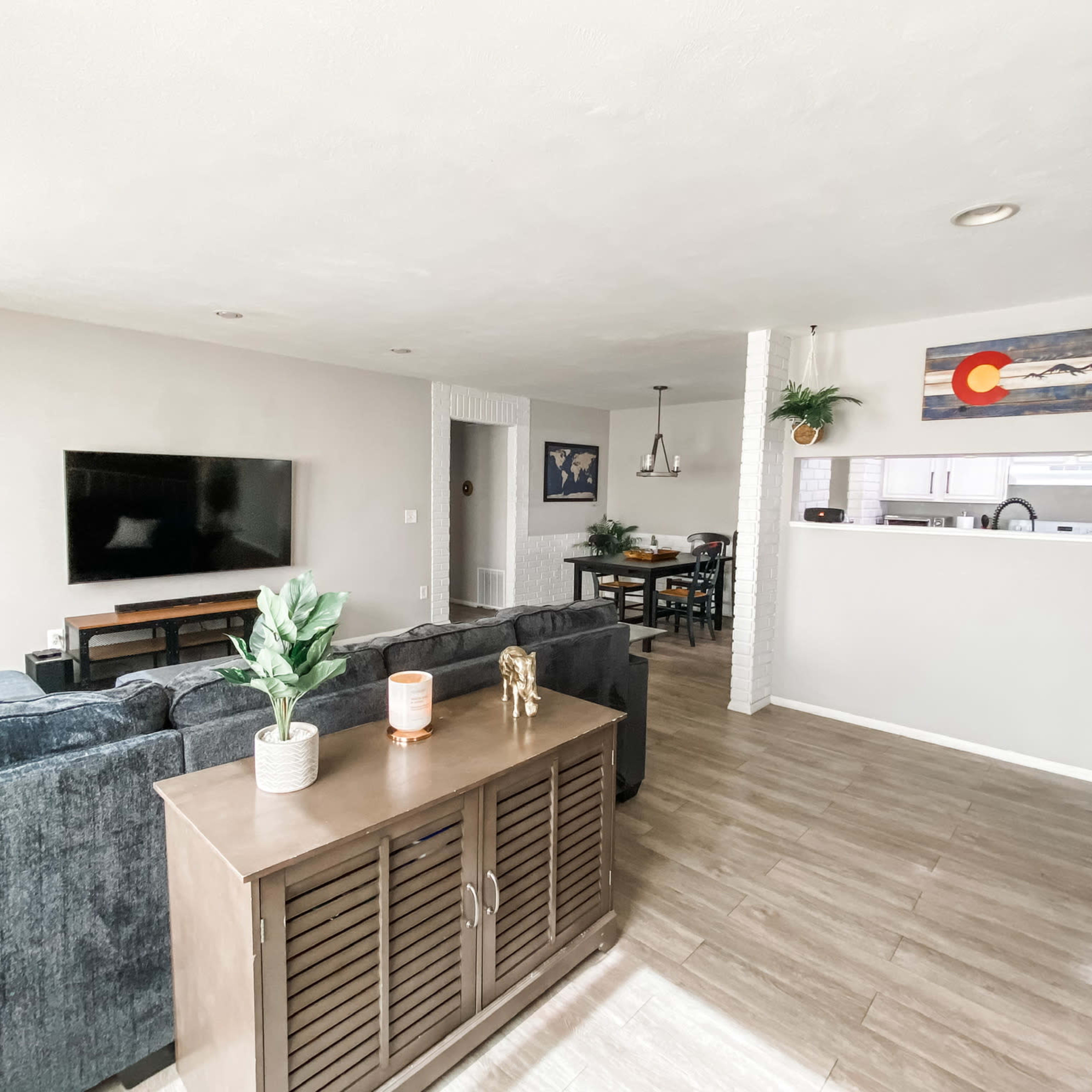 The living room features a dark couch facing a wall-mounted television, with a dining area visible in the background that includes a table and chairs.