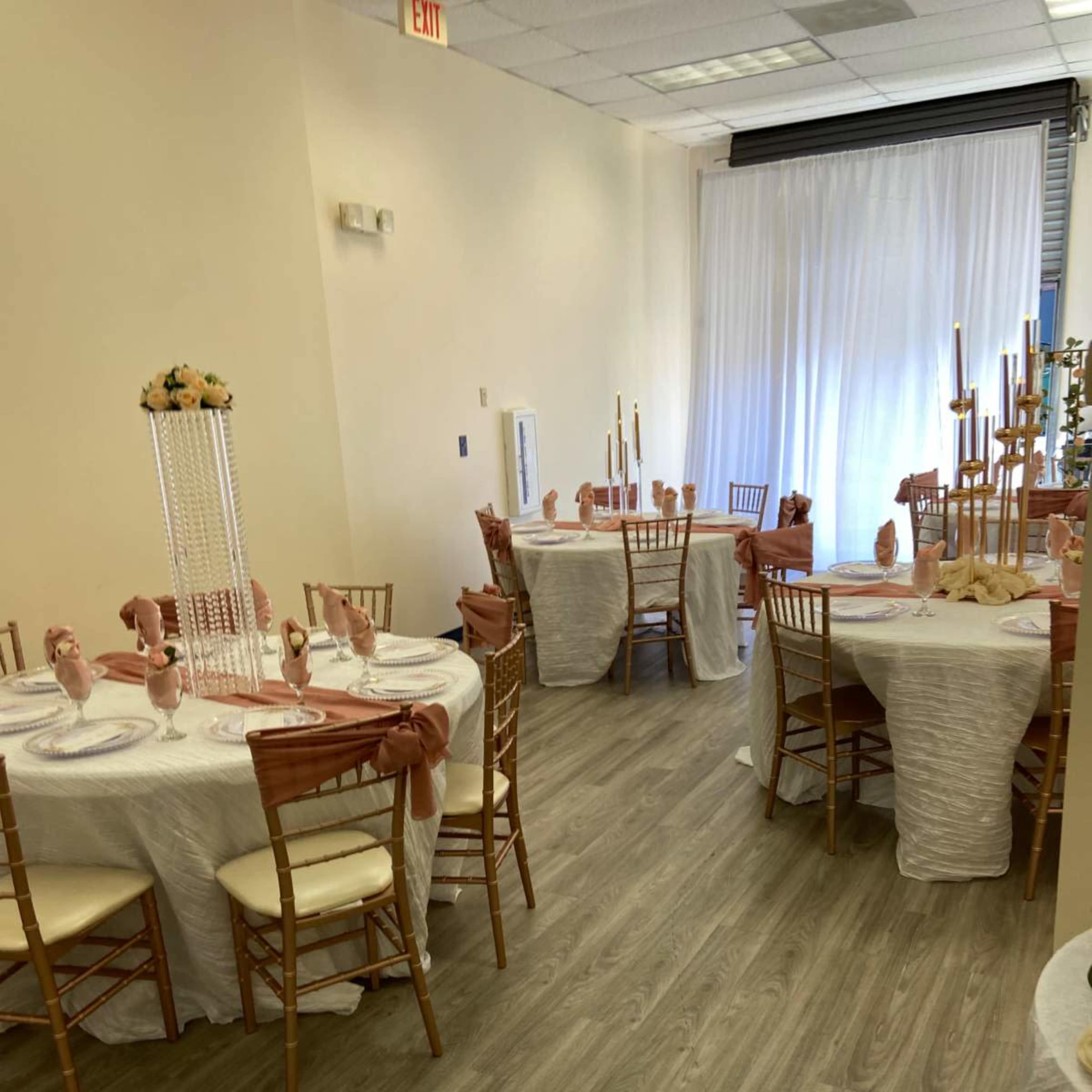 A decorated dining area with round tables set with white tablecloths and golden chairs, illuminated by natural light from a large window.