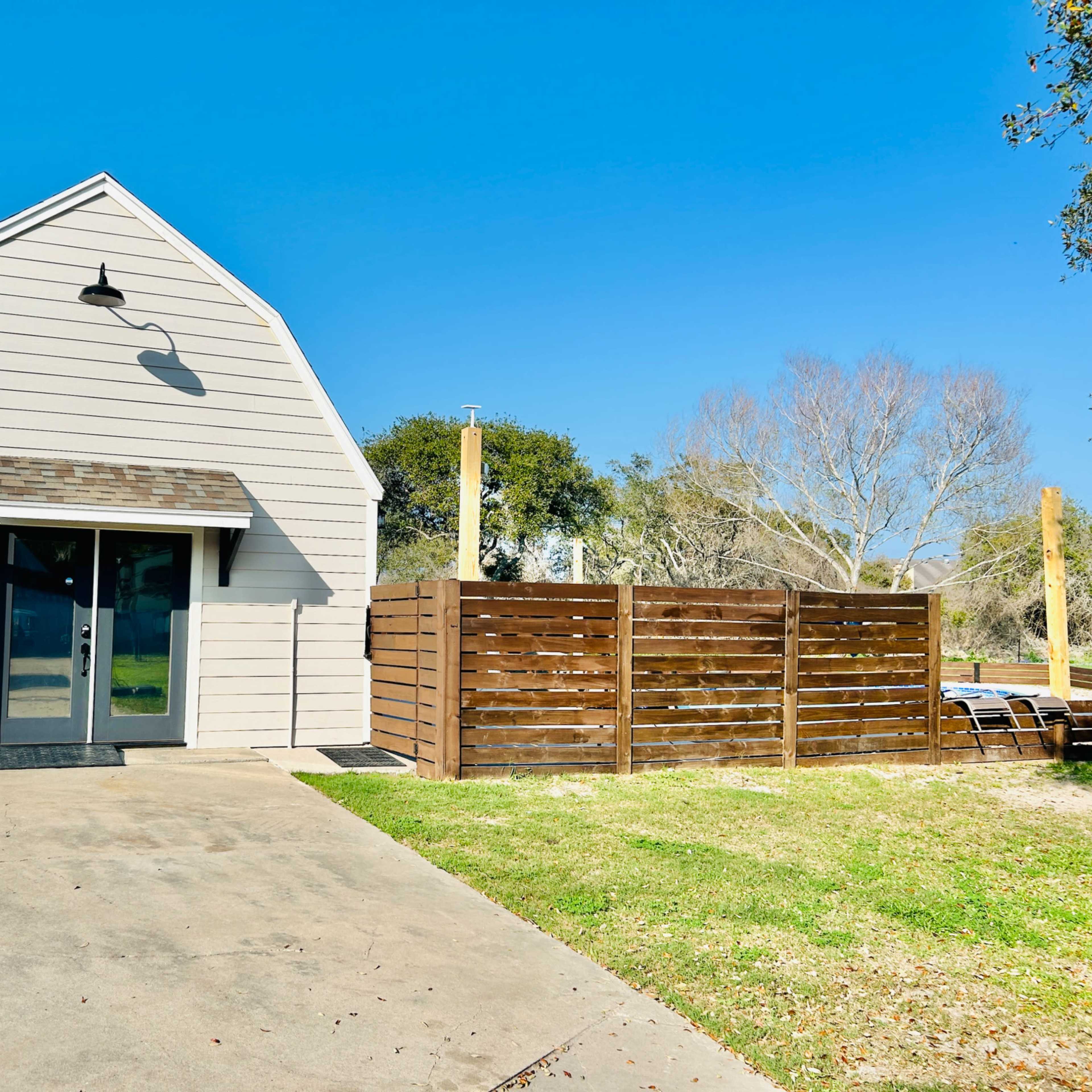A small, gabled house with a paved driveway is situated beside a fenced yard against a clear blue sky.