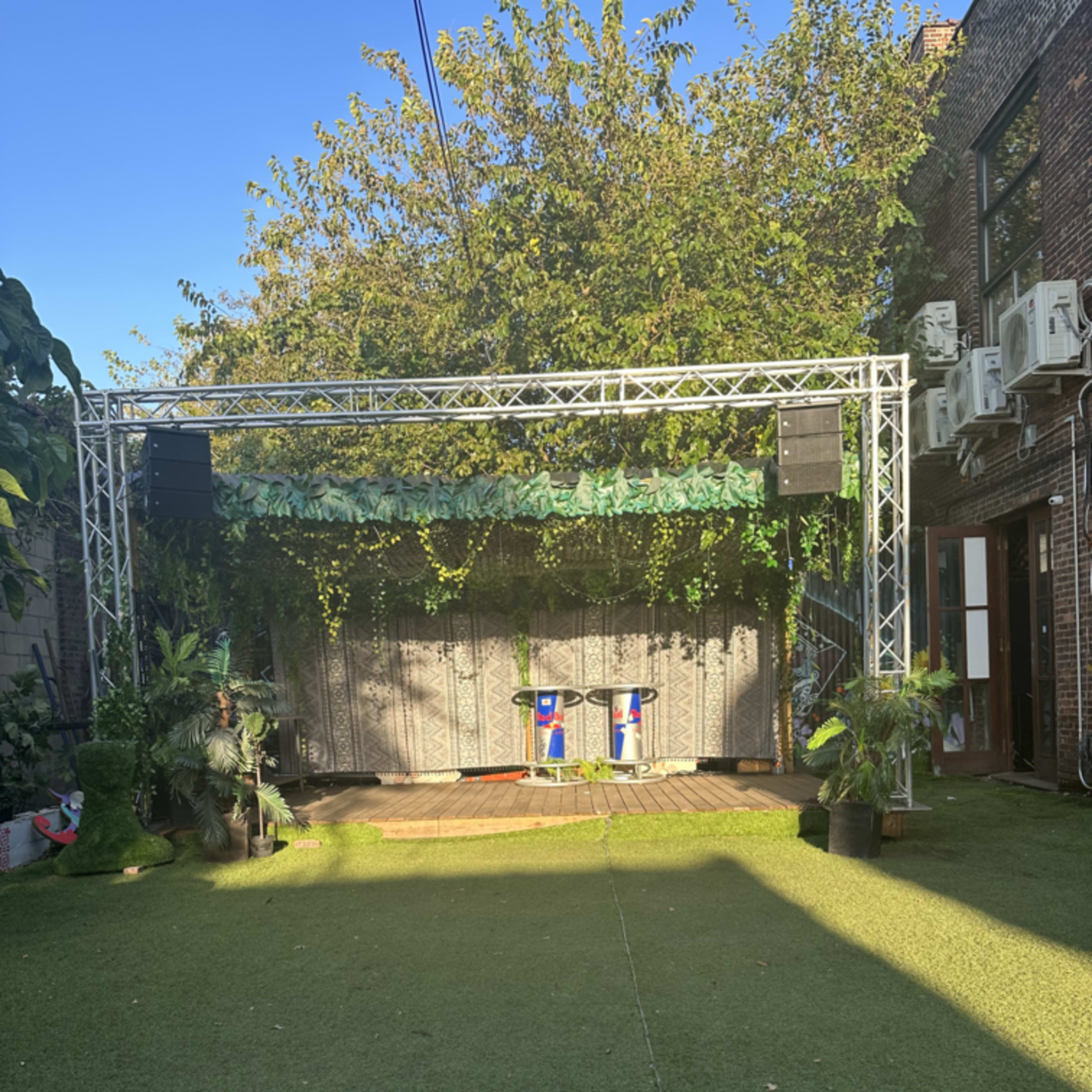 An outdoor stage setup with a greenery backdrop, flanked by speakers and surrounded by potted plants on a grassy surface.