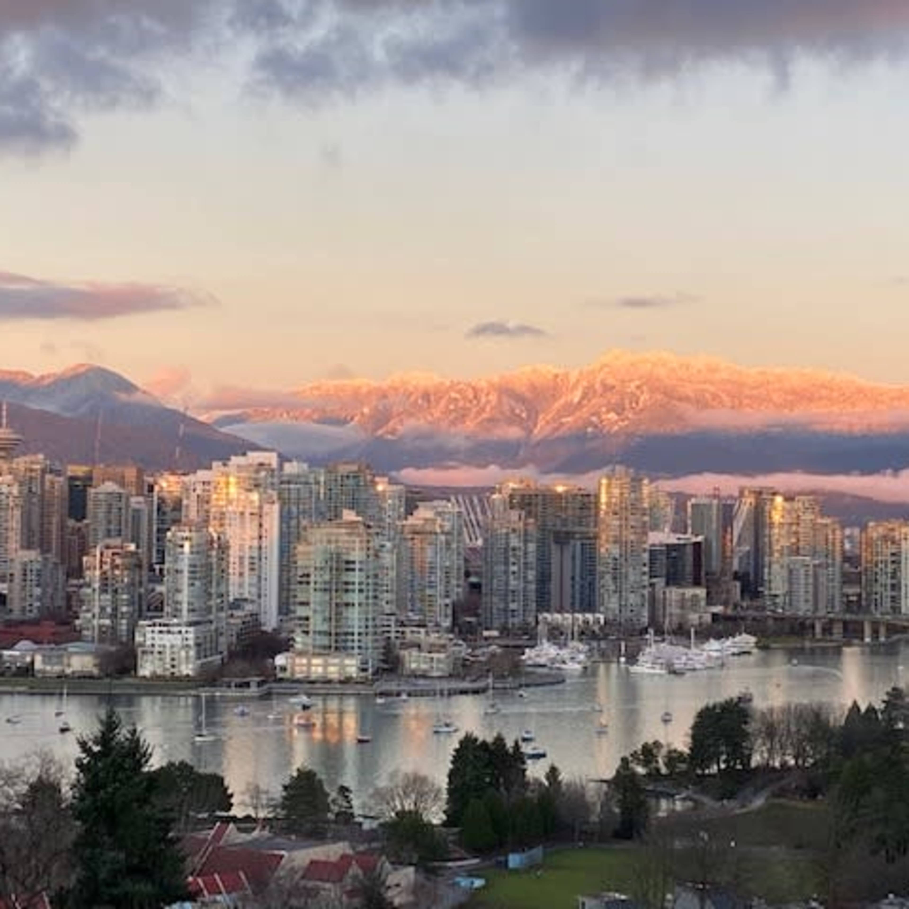A city skyline with tall buildings beside a river, set against mountains with sunlight illuminating their peaks.