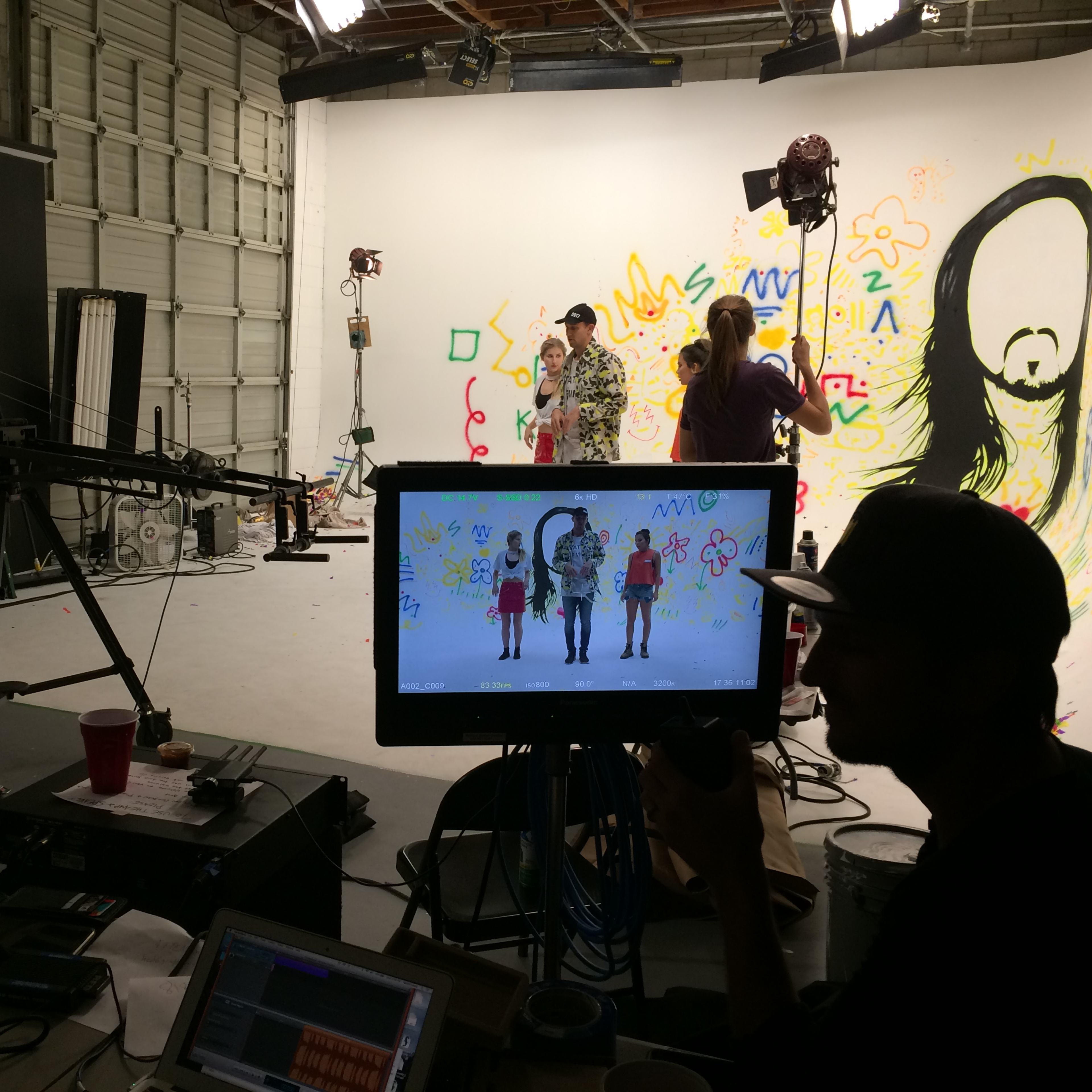 A film crew works on a set with brightly painted backgrounds while monitoring the scene on a screen.