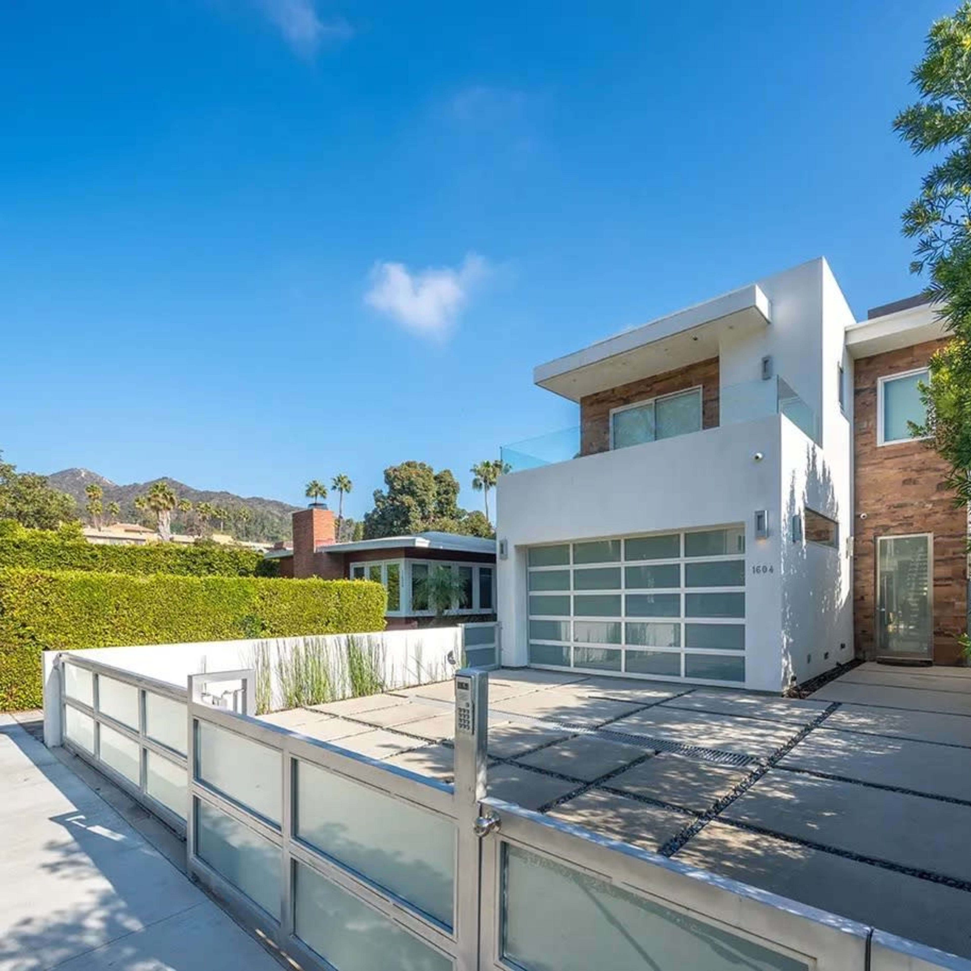 A modern two-story house with a flat roof, large windows, and a glass garage door, surrounded by neatly trimmed hedges and a paved driveway.