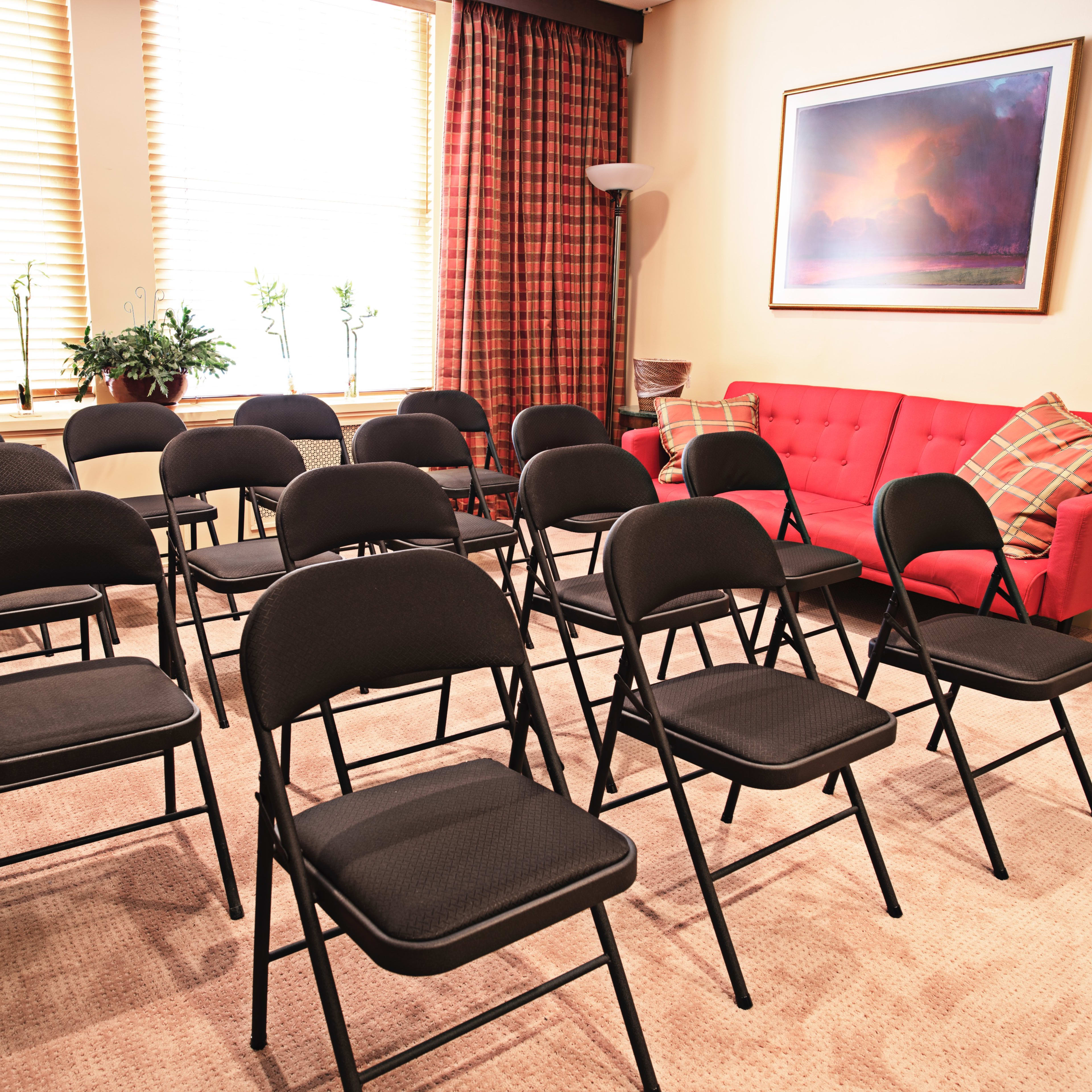A small room features a set of black folding chairs arranged in rows facing a red couch and large windows with light filtering through the curtains.