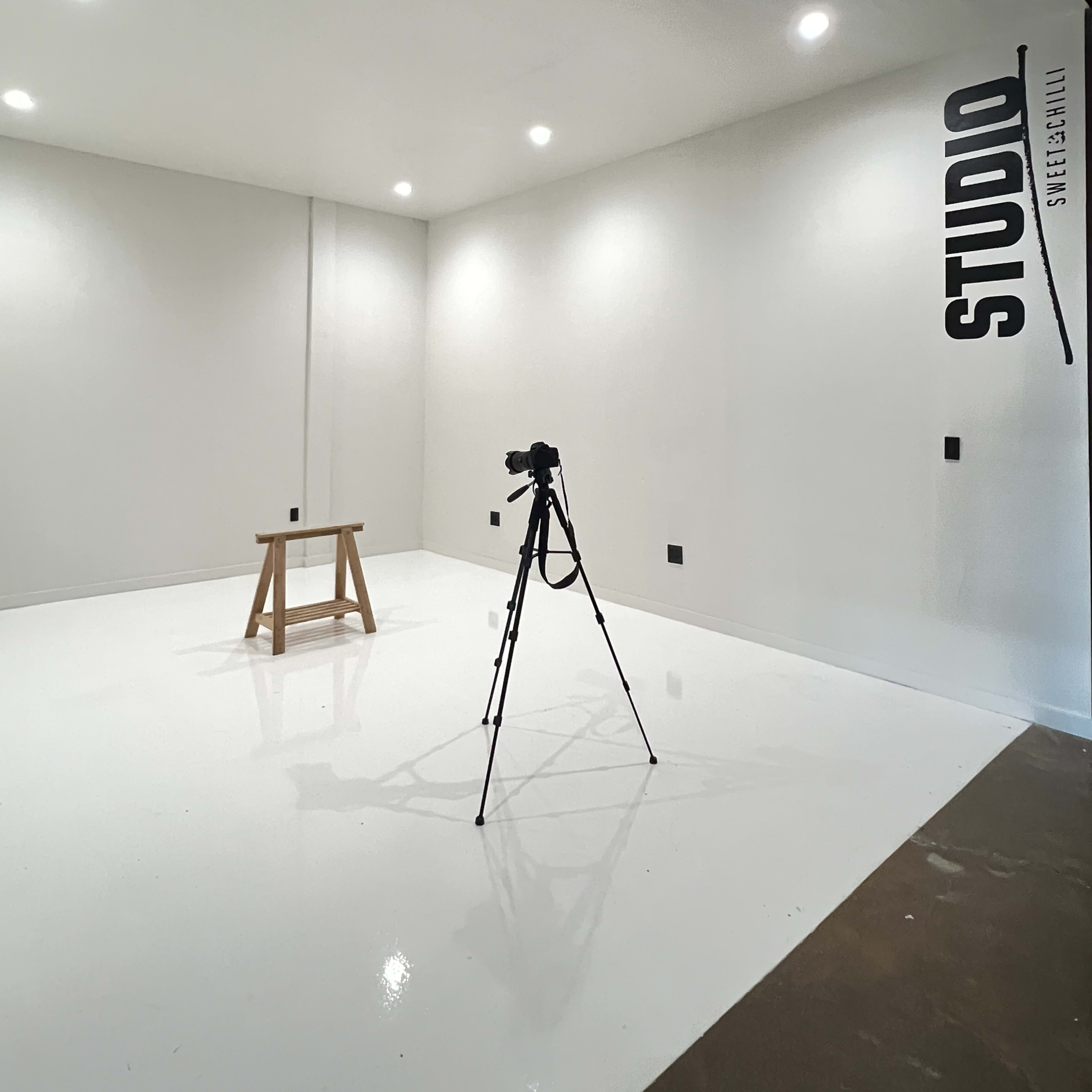 A minimalistic studio space with a wooden stool and a camera tripod positioned on a white floor under bright lights.