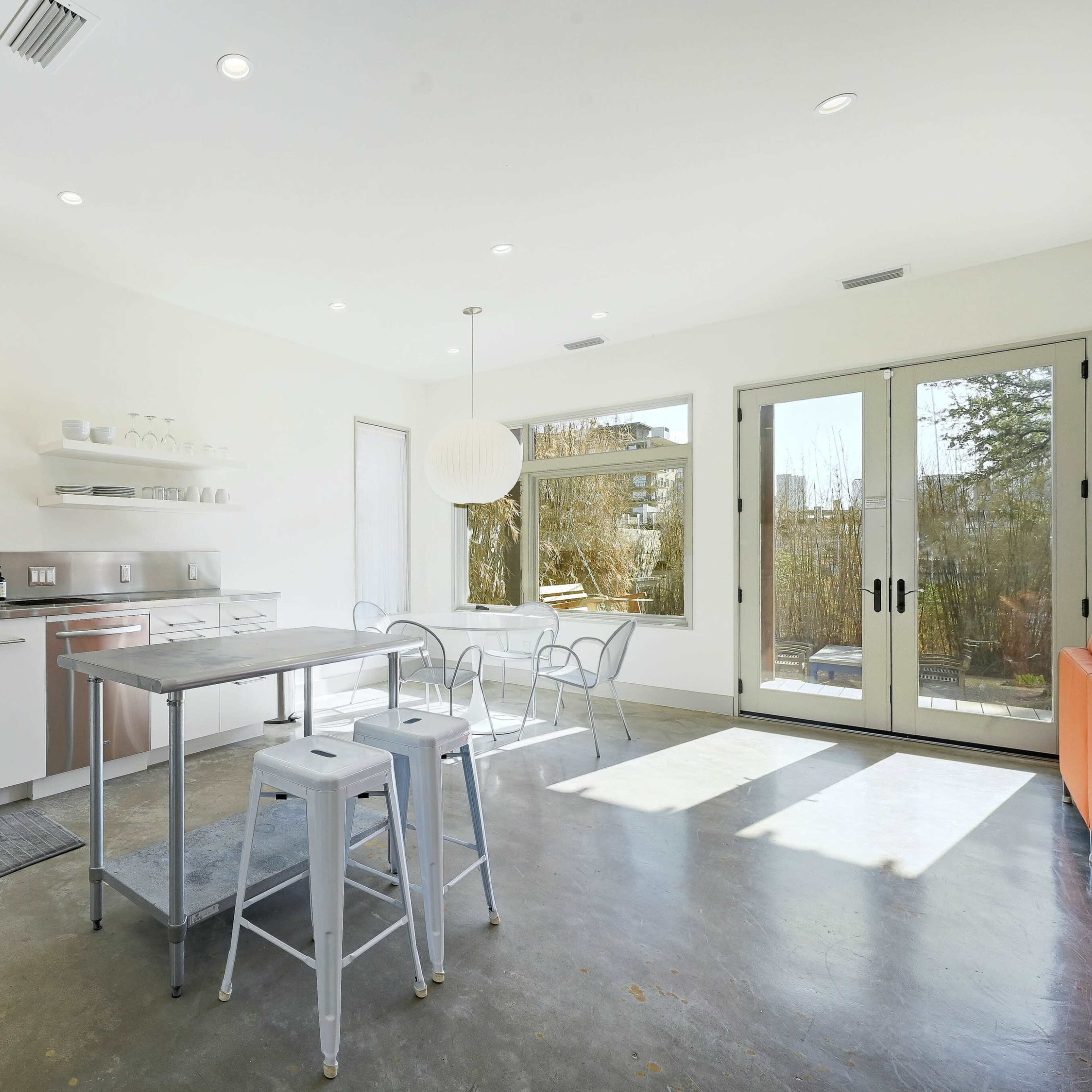 A modern kitchen with stainless steel appliances, white cabinetry, a central table with metal stools, and large windows overlooking a natural setting.