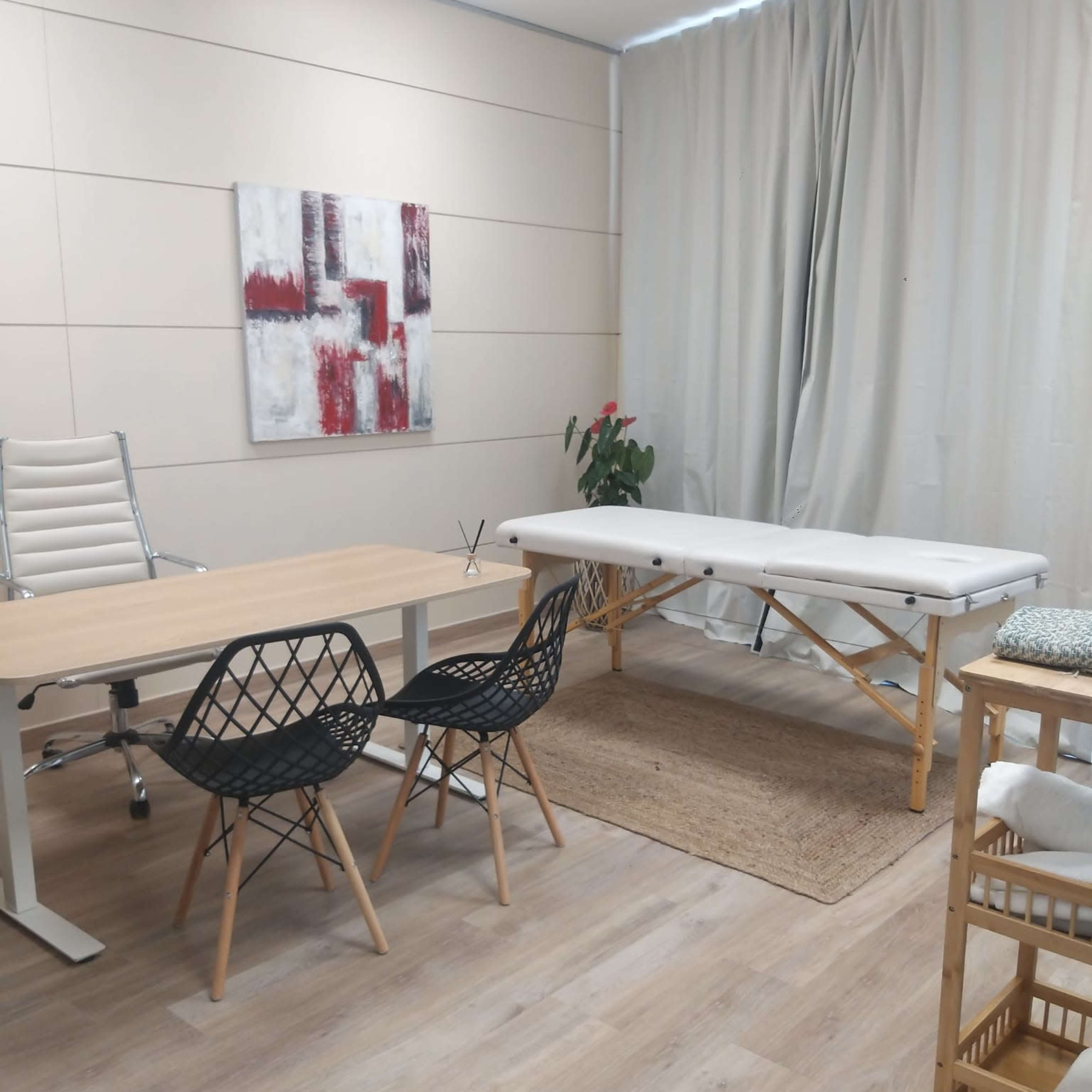 A small, well-organized room features a wooden table with two black chairs, a massage table, and a plant, with light-colored walls and curtains.