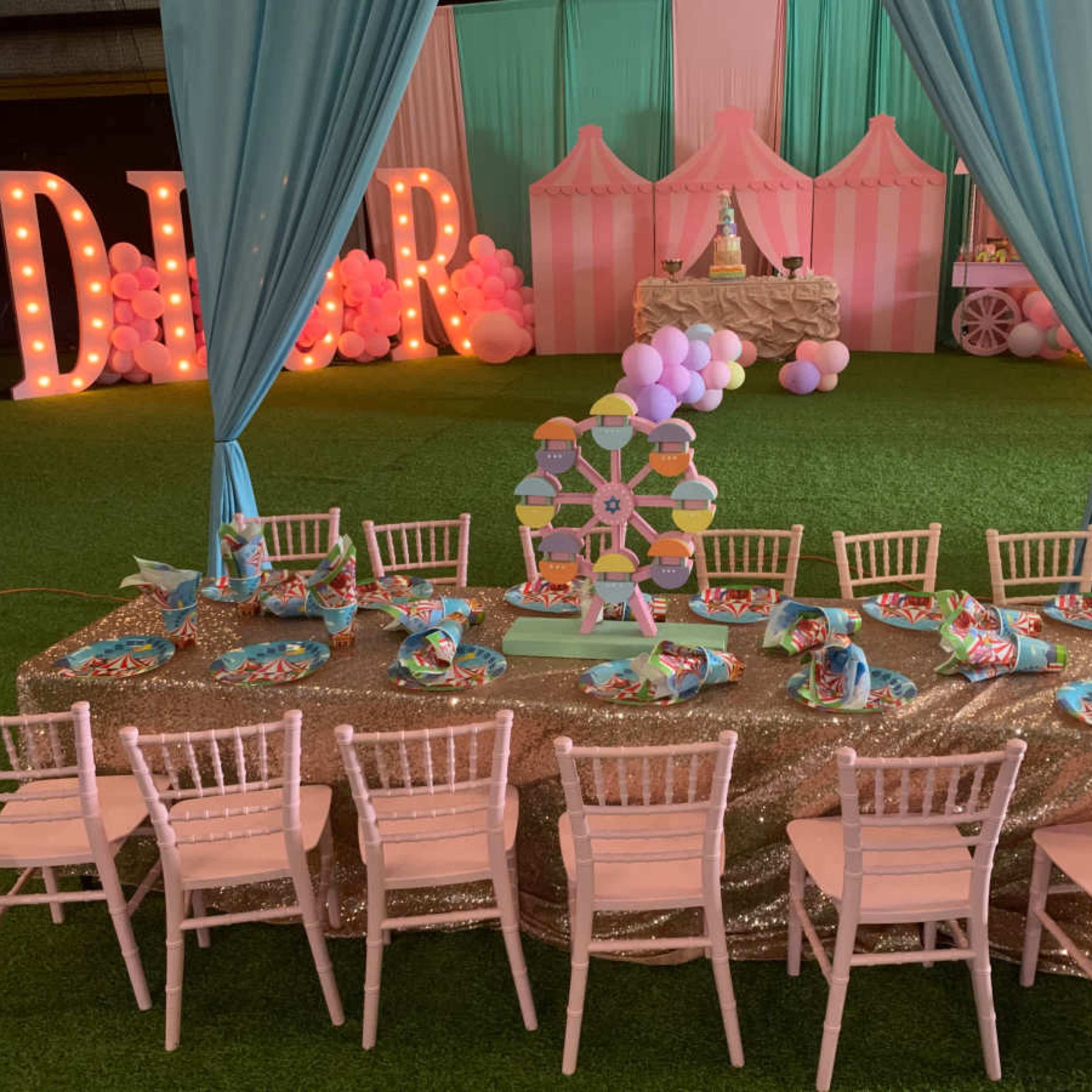A decorated event space features a long table set with colorful plates and a centerpiece, surrounded by pink chairs under draped fabric and balloons.