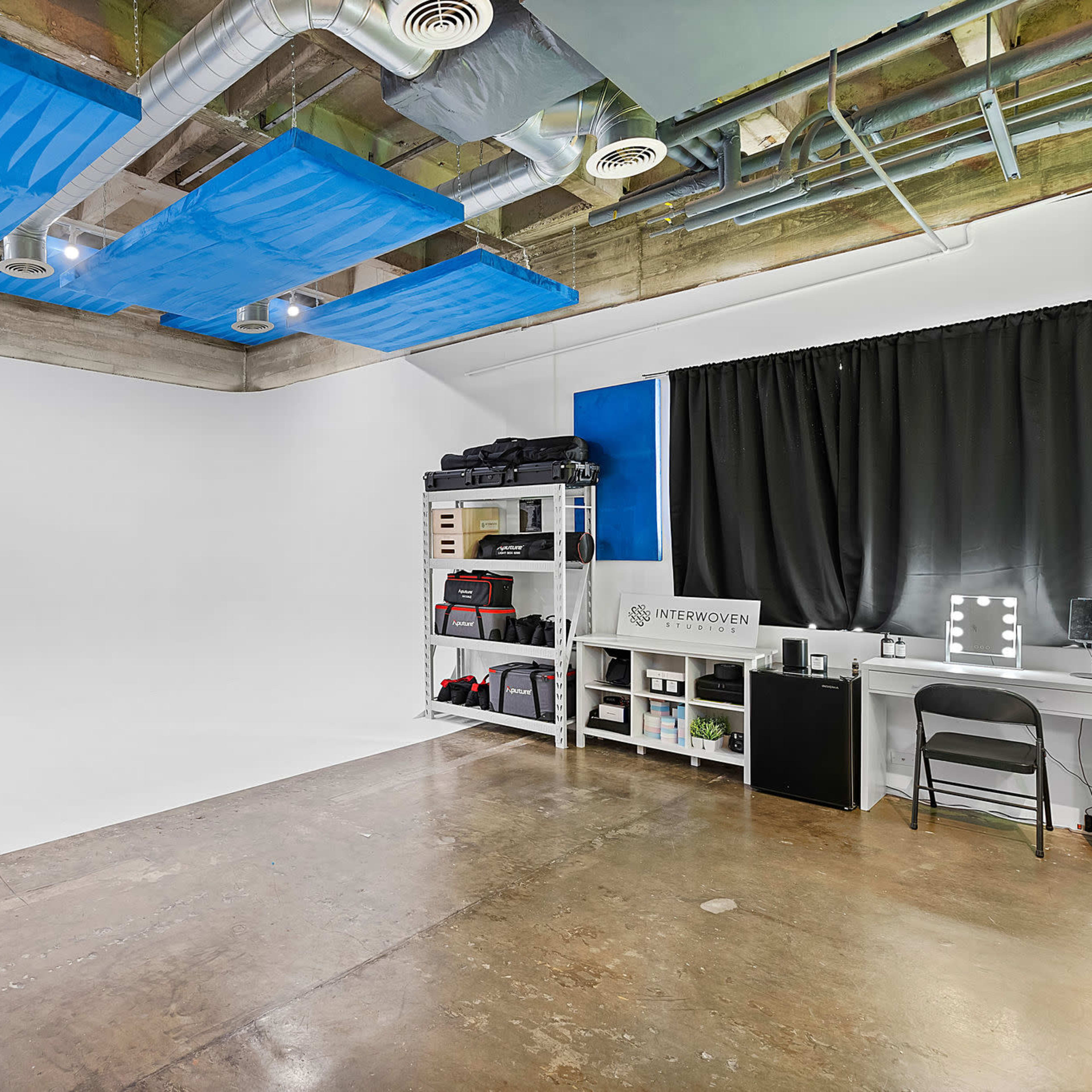A minimalistic studio space featuring a white backdrop, concrete flooring, and various equipment arranged along the wall.