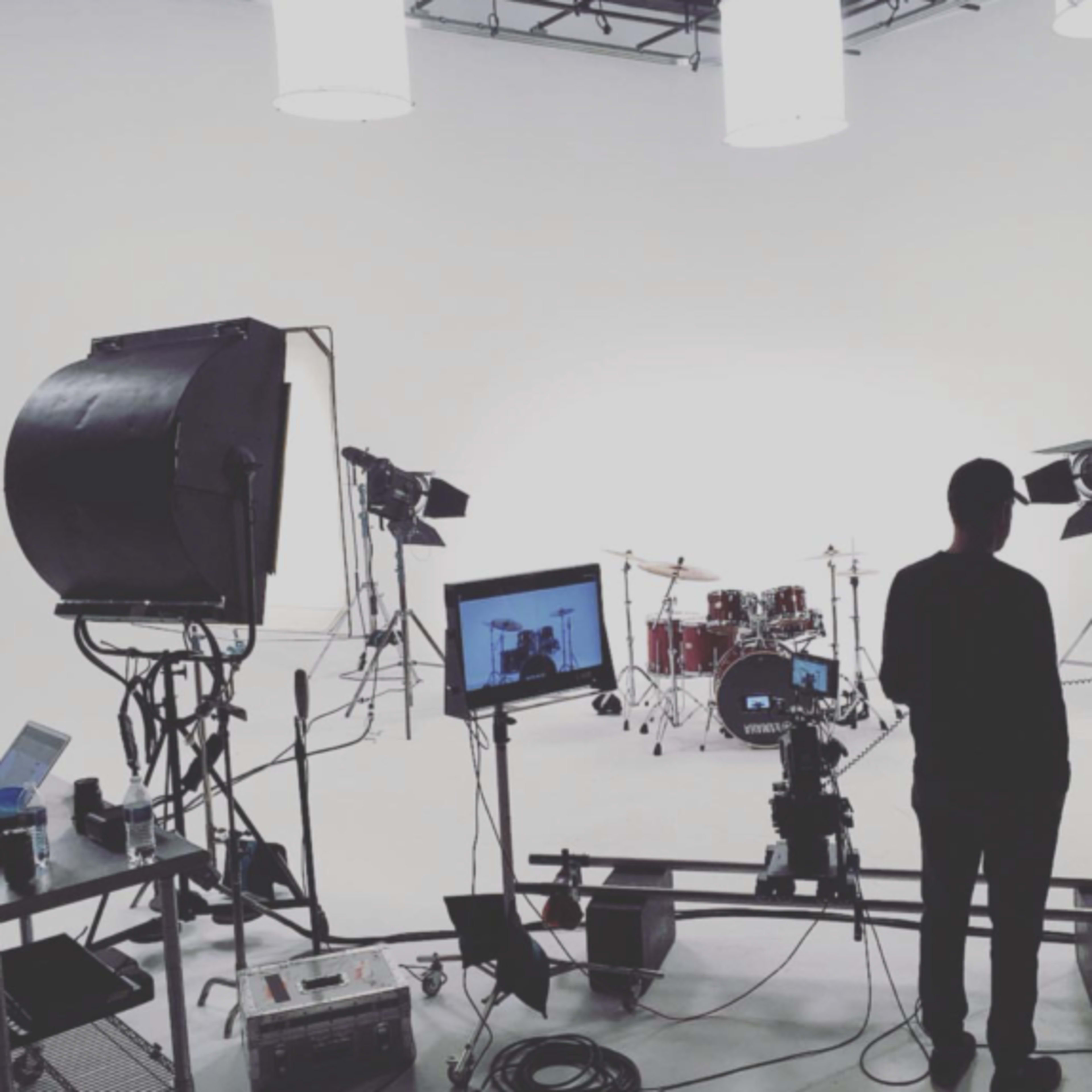 A person stands in a studio surrounded by camera equipment and lighting, with a drum set positioned in the background.