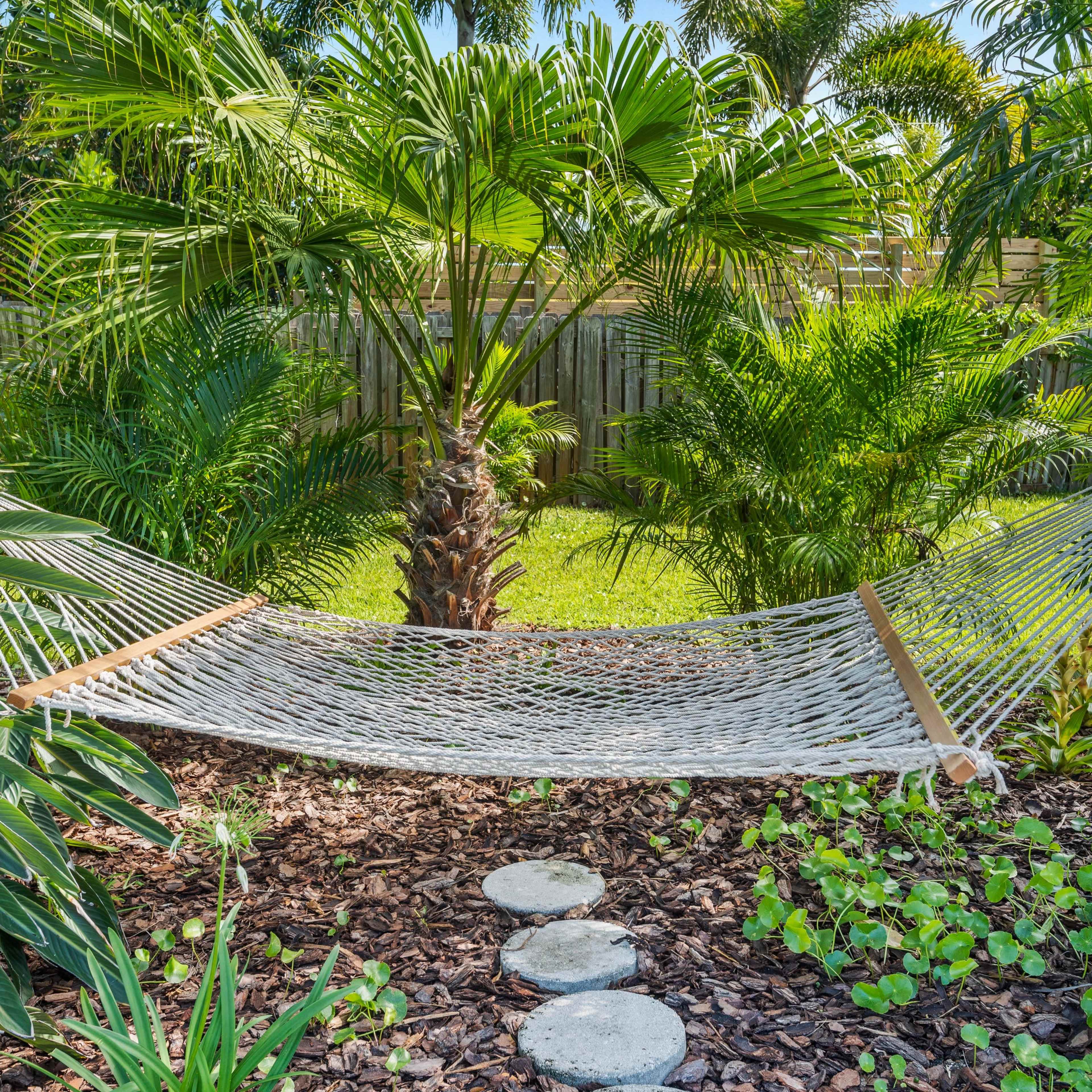 A hammock is suspended between two palm trees in a lush, green garden, surrounded by various plants and a stone pathway.