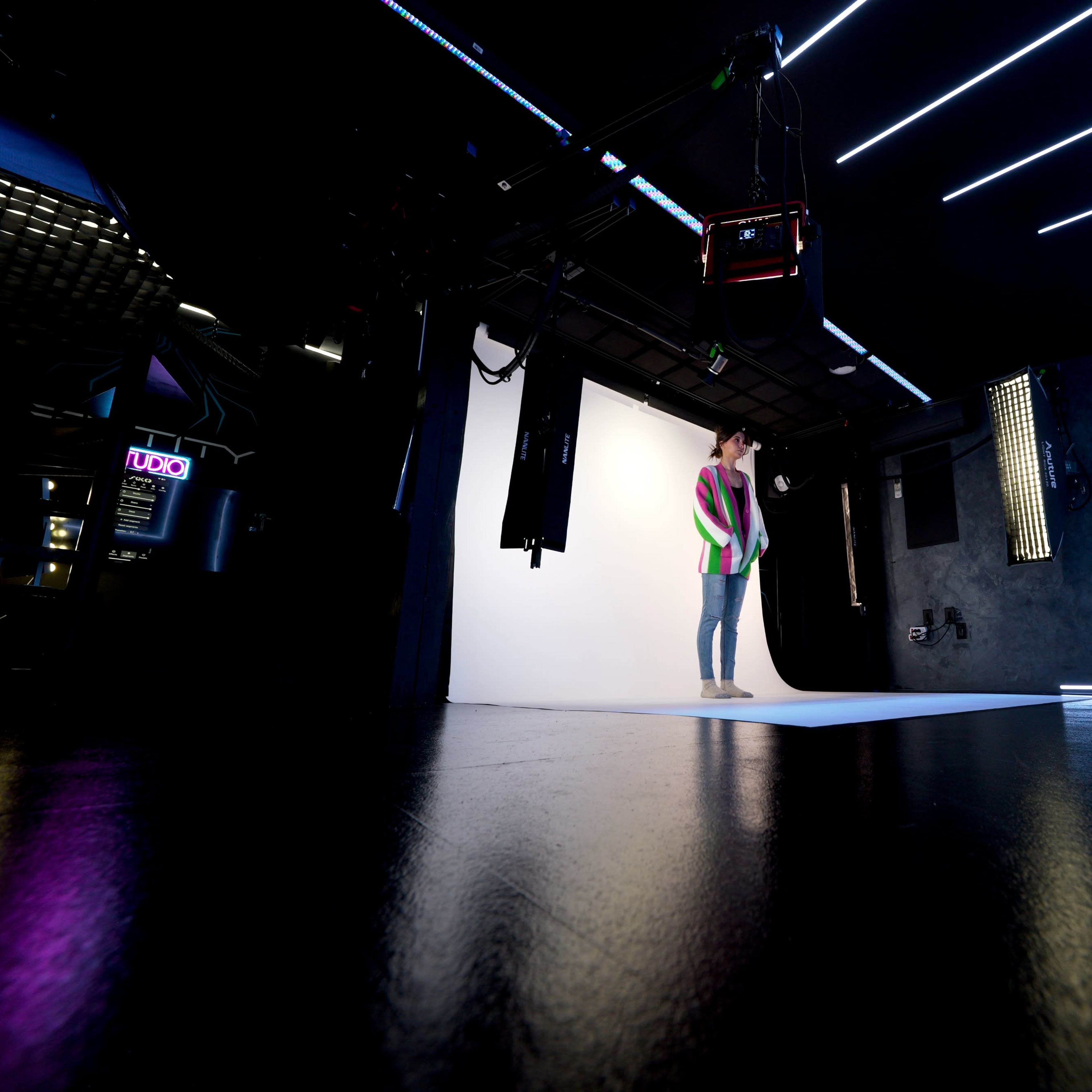 A person poses on a photo backdrop while another person sets up a camera in a well-lit studio with contrasting neon lights.
