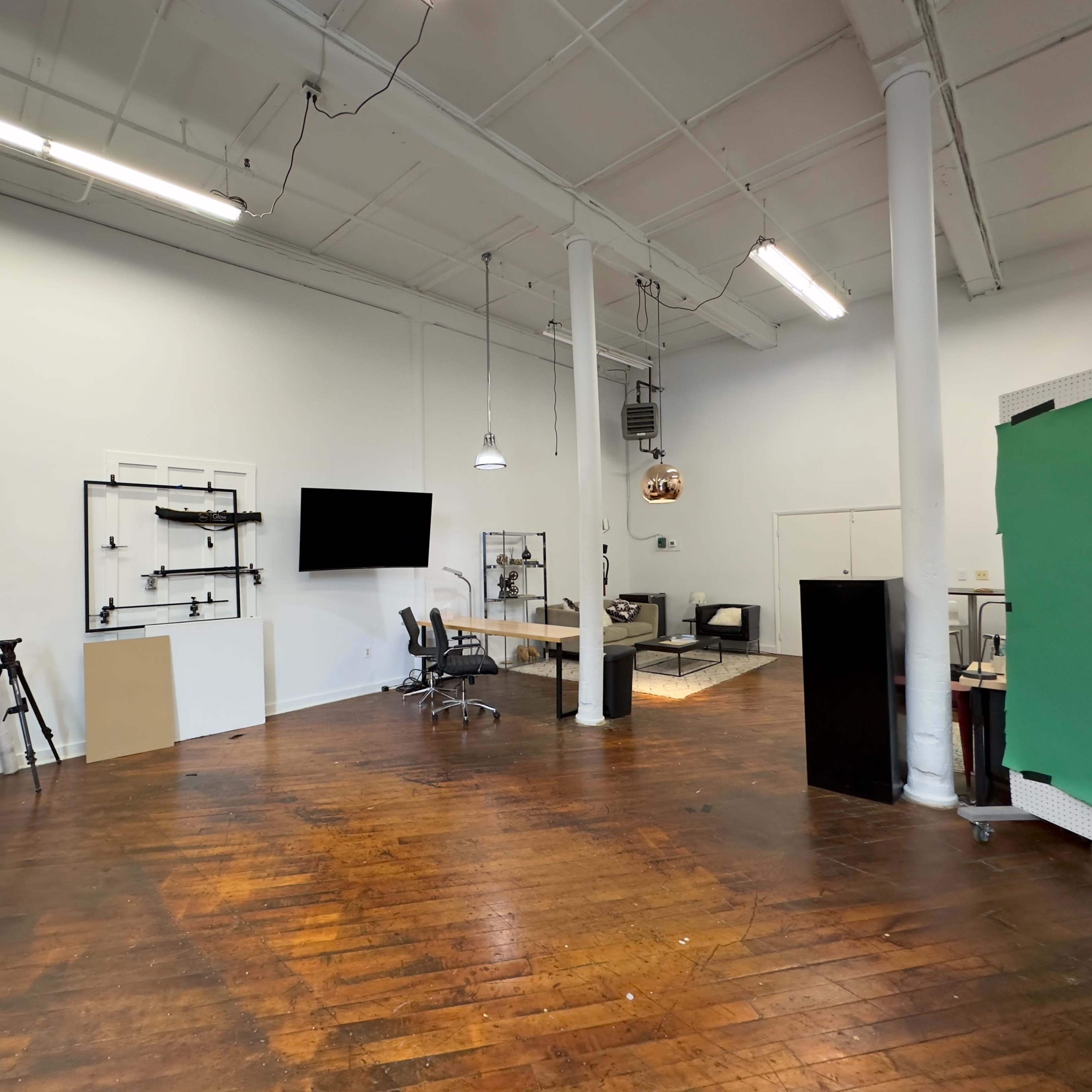 A spacious, well-lit studio with wooden floors, a green backdrop, shelves holding equipment, and a workspace featuring a large table and chairs.