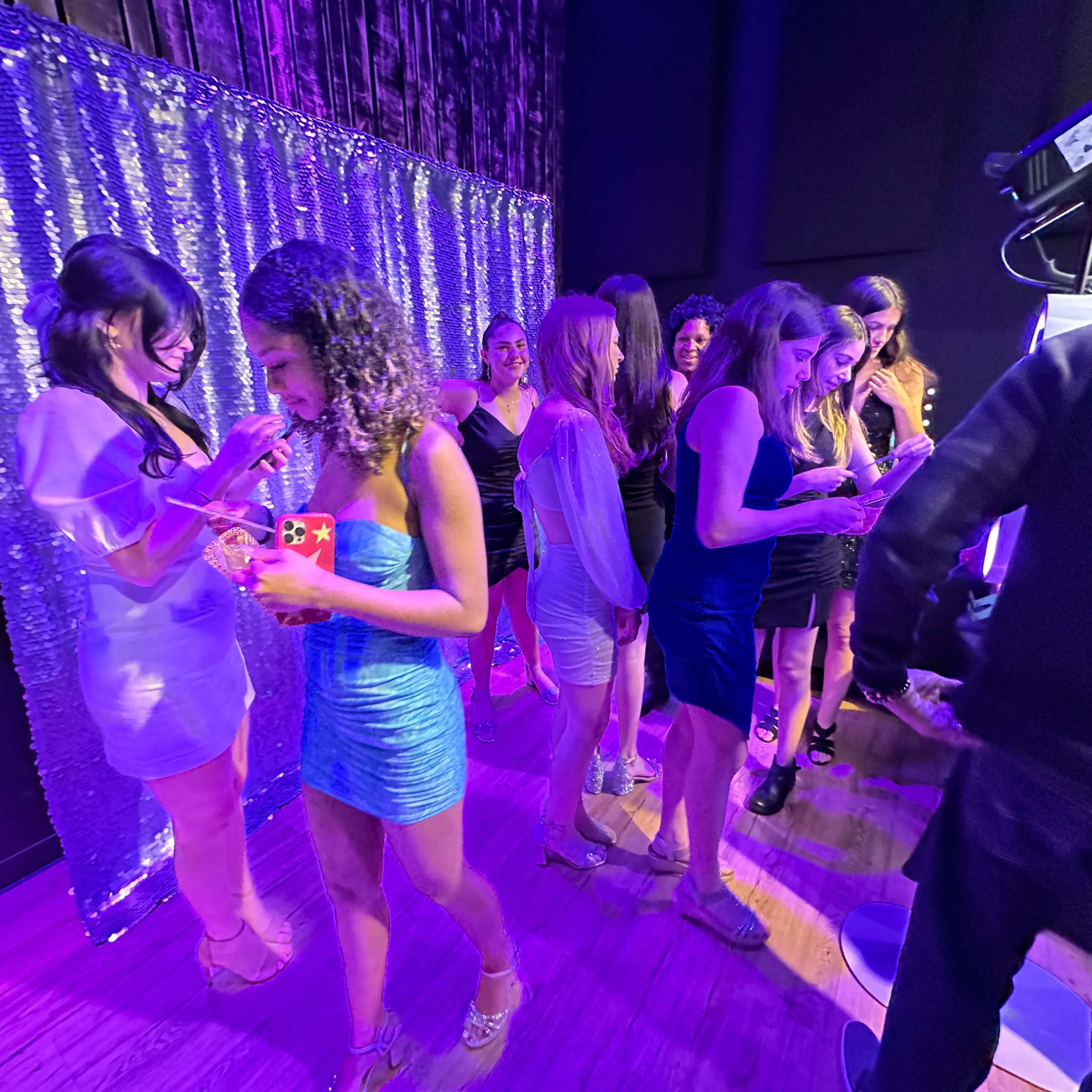A group of women dressed in evening attire stands in front of a shimmering backdrop at a social event.