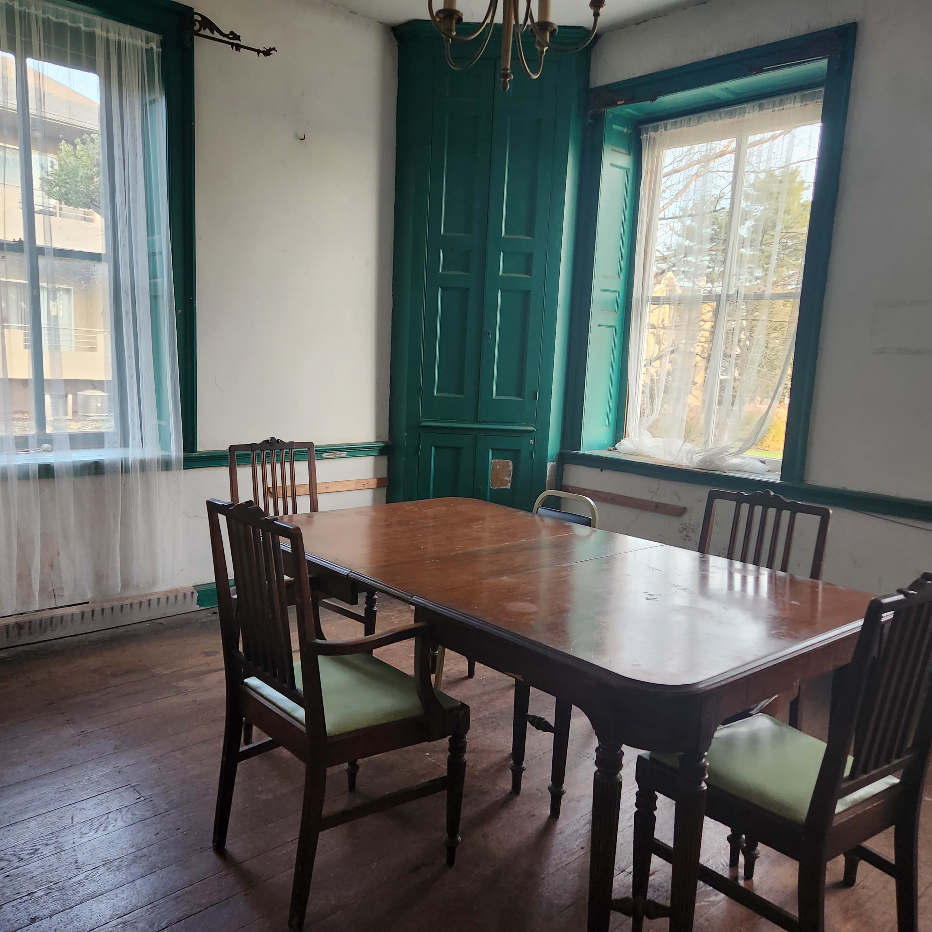 A wooden table surrounded by four chairs is positioned in a room with green accents and large windows.