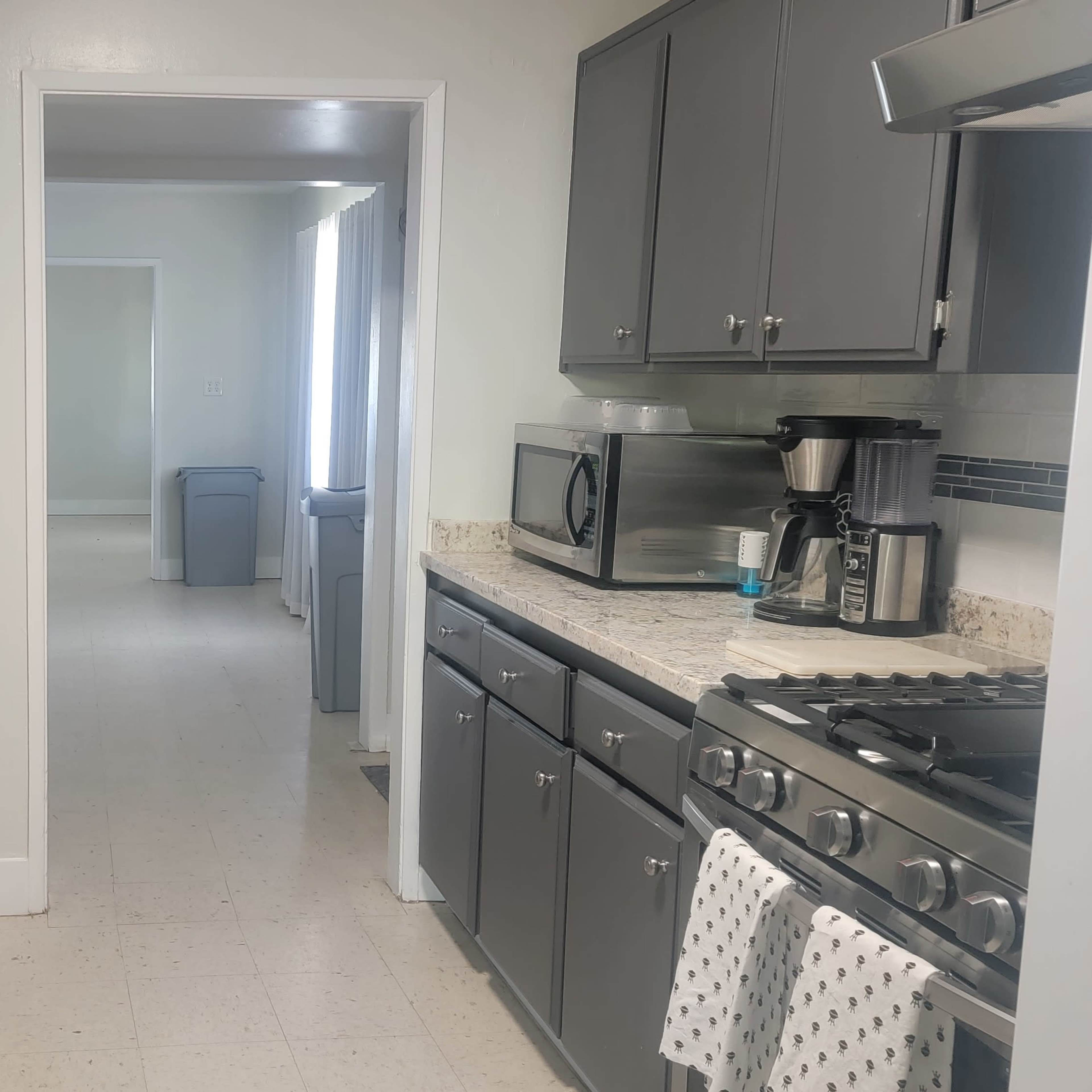 A gray-themed kitchen with granite countertops, featuring a sink, stove, and various appliances, leading into a hallway with a visible laundry area.