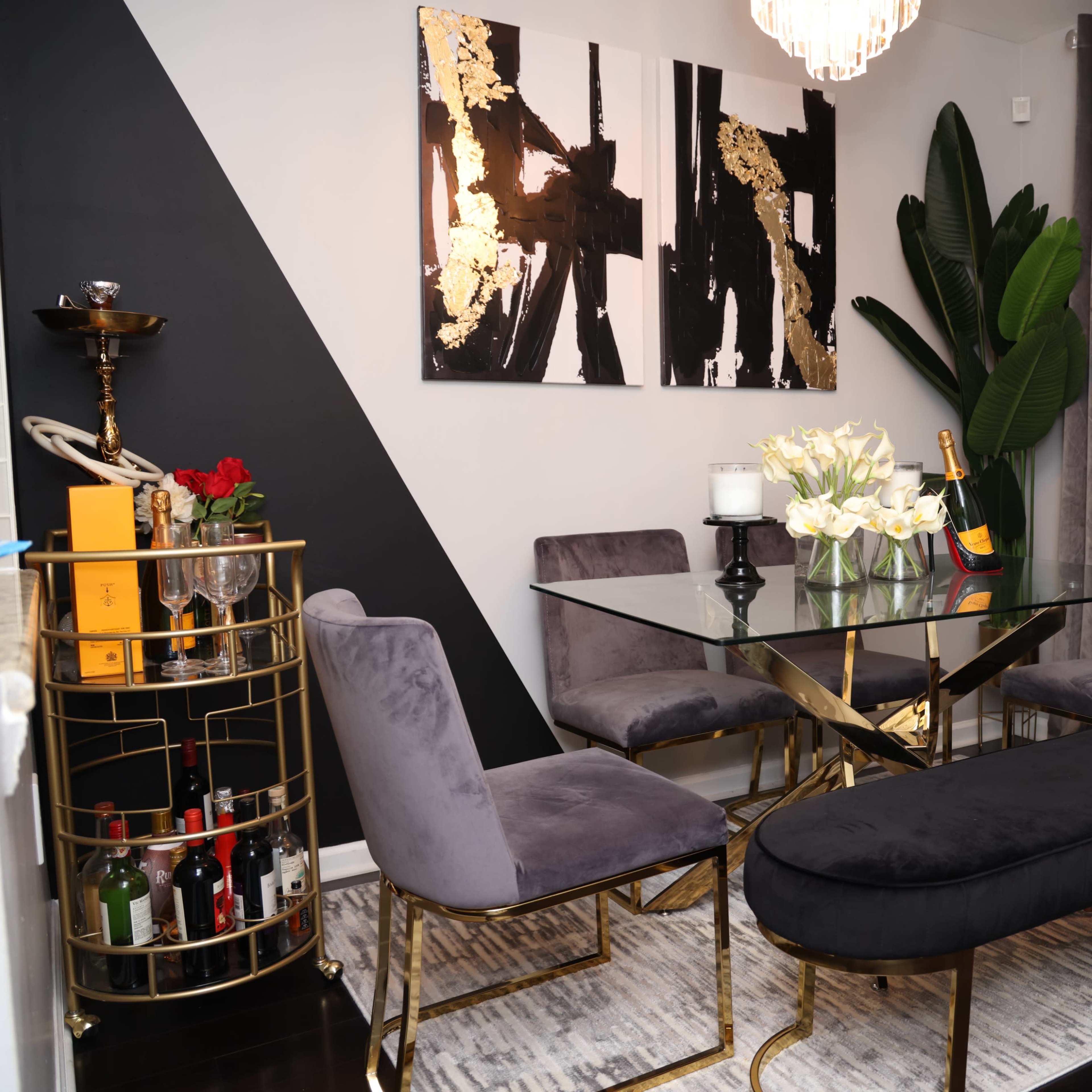 A modern dining area features a glass table surrounded by gray velvet chairs, a gold-accented bench, and a bar cart filled with bottles against a wall adorned with abstract art.