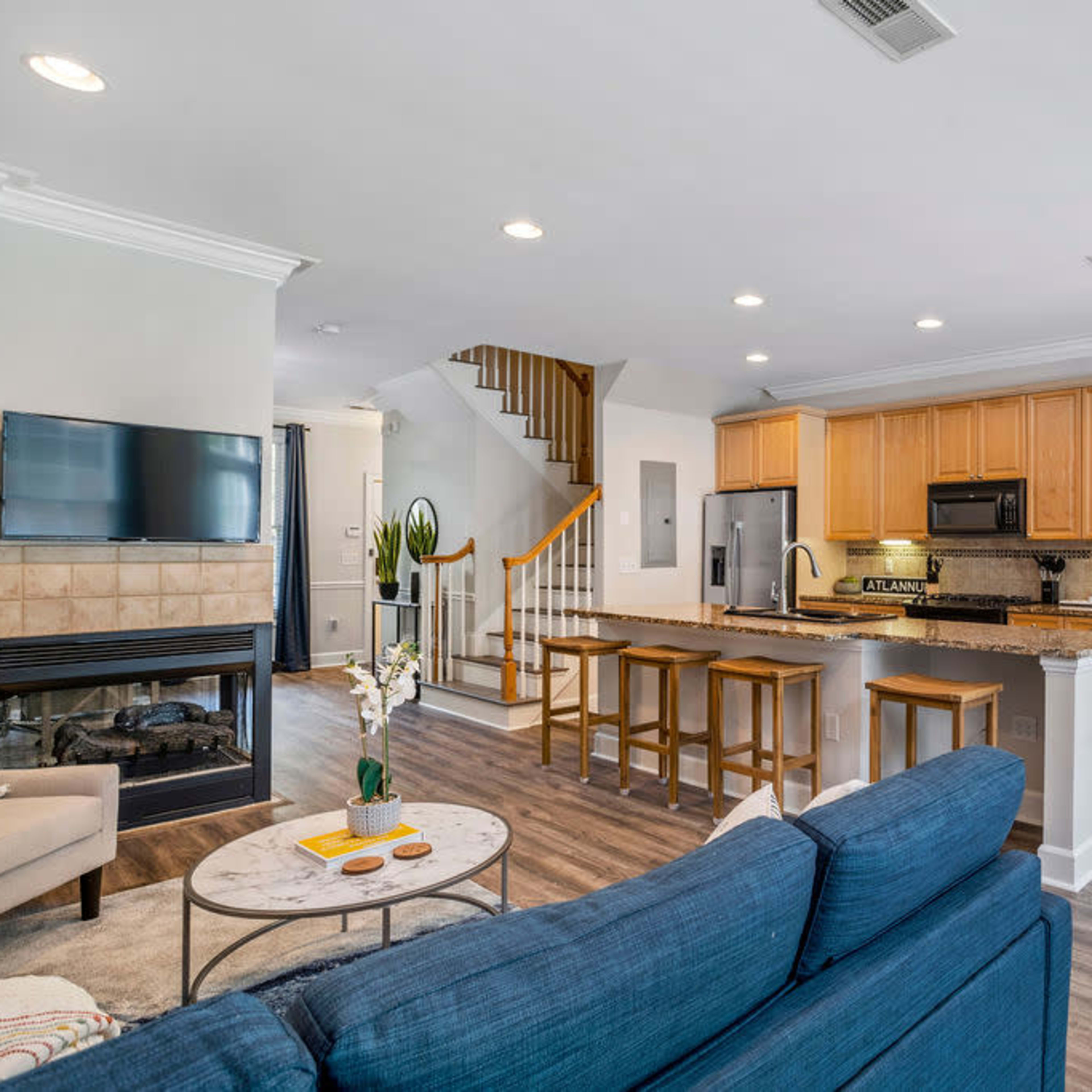 A modern living space featuring a kitchen with wooden cabinets, a dining area, a living room with a blue sofa, and a staircase leading to an upper level.