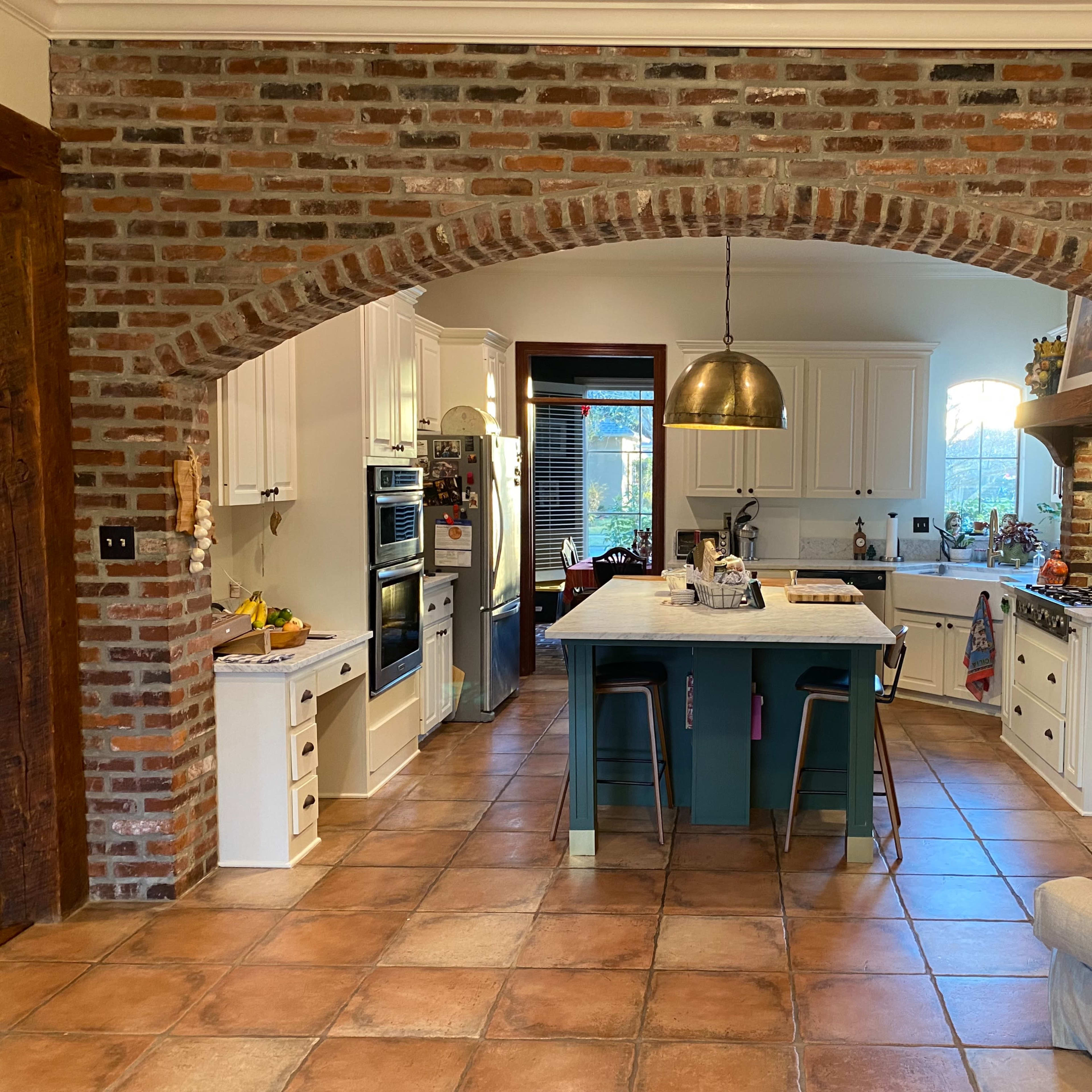 A kitchen with a brick archway, featuring white cabinetry, a large island with bar stools, and tiled flooring.