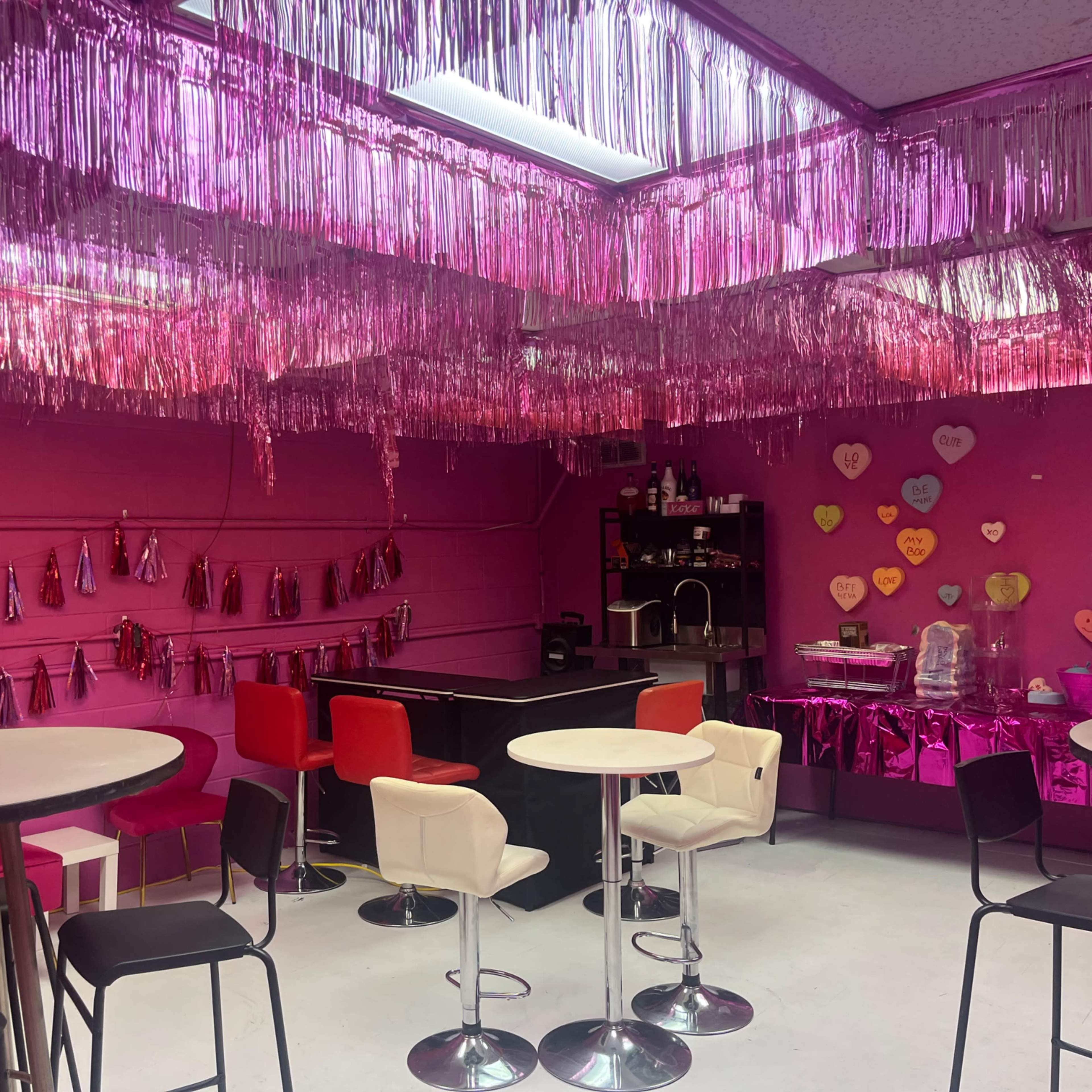The room features pink walls and ceiling decorations with hanging metallic strands, a bar area, and several tables and chairs arranged for a social setting.