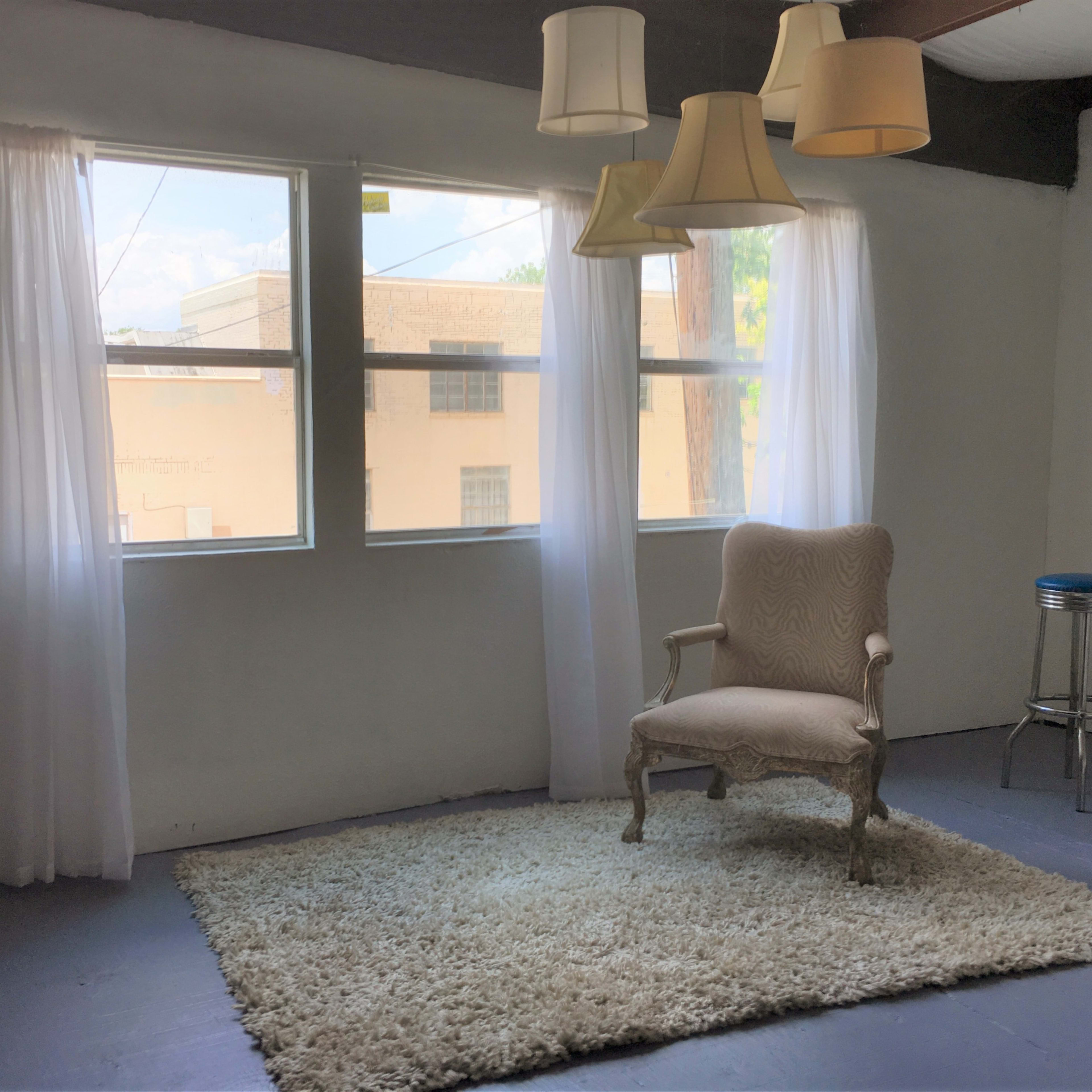 A decorated room features a plush rug, a vintage chair, and hanging light fixtures near large windows with sheer curtains.