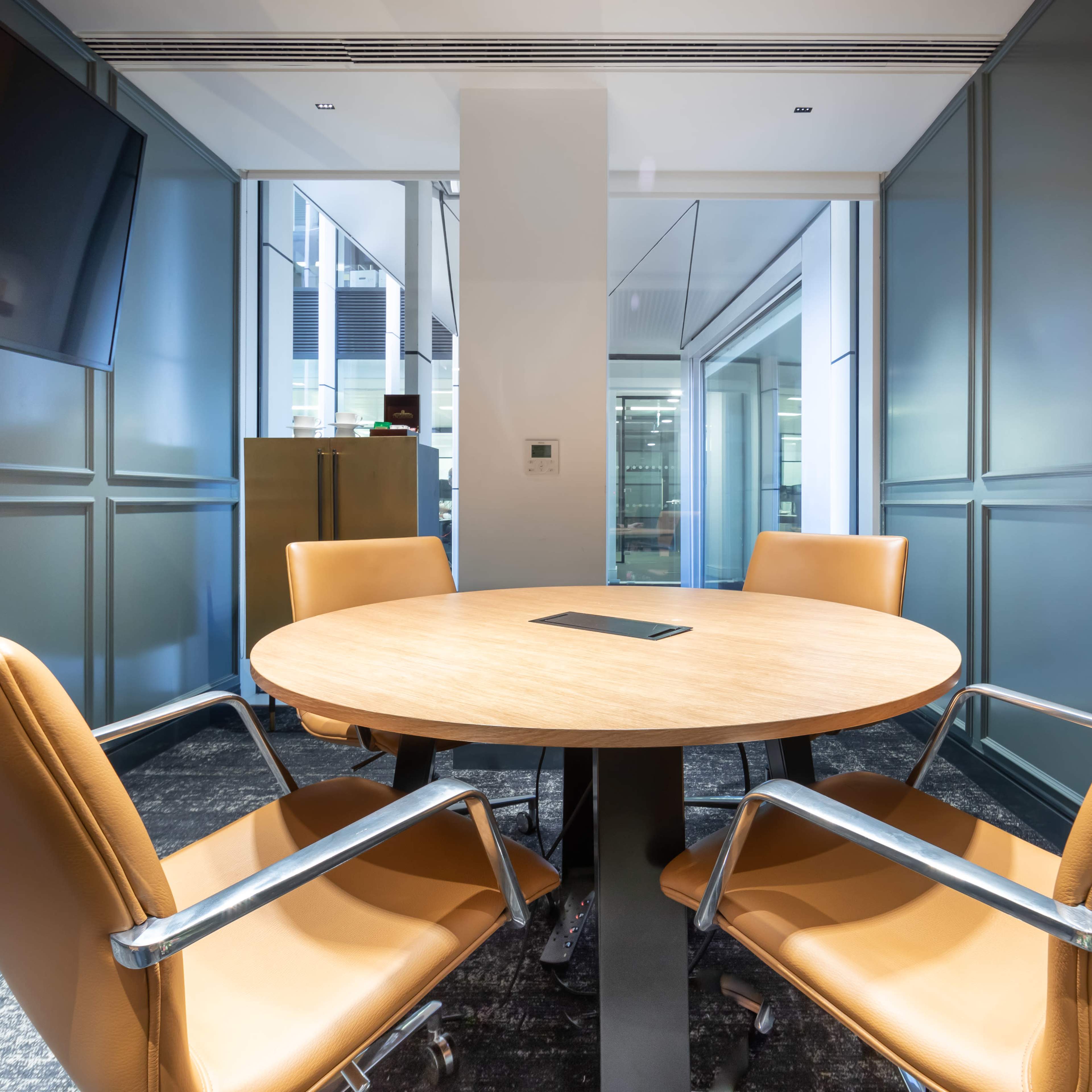 A modern conference room with a round wooden table and four leather chairs arranged around it, against a backdrop of glass walls.