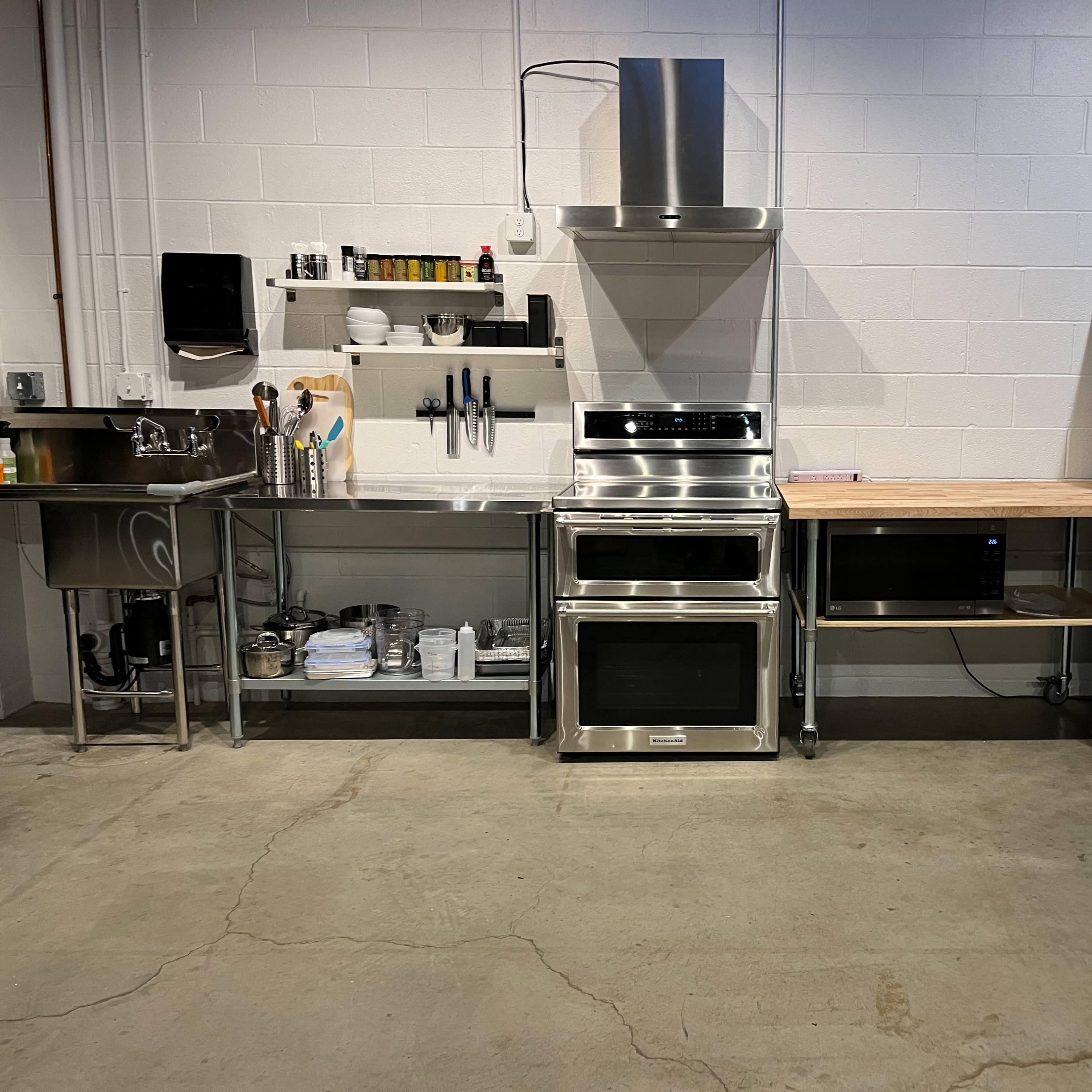A modern kitchen featuring a stainless steel cooking range, an oven, a stainless steel sink with shelving, a countertop, and a large refrigerator against a concrete wall.