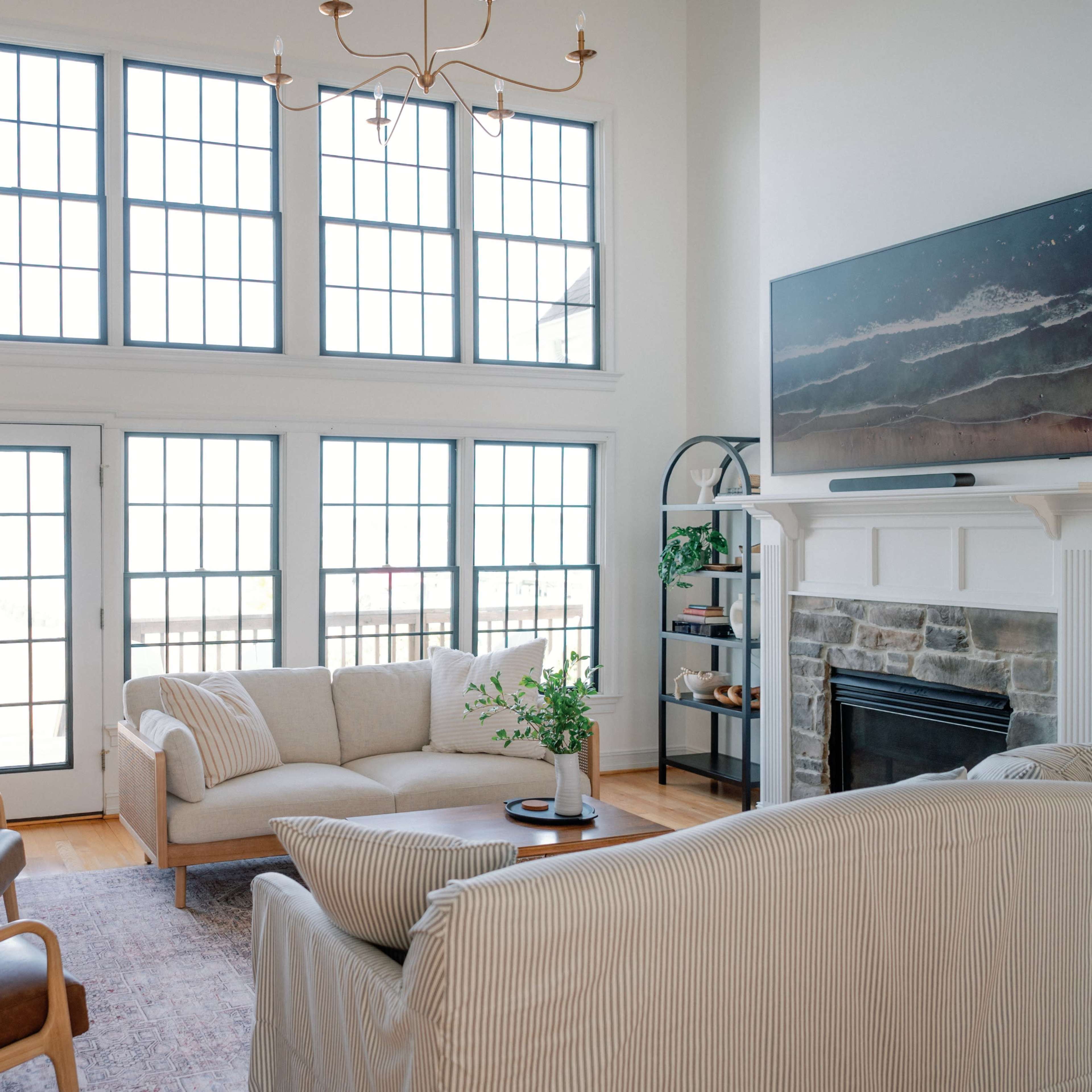 A spacious living room features large windows, a light-colored sofa, two armchairs, and a stone fireplace.
