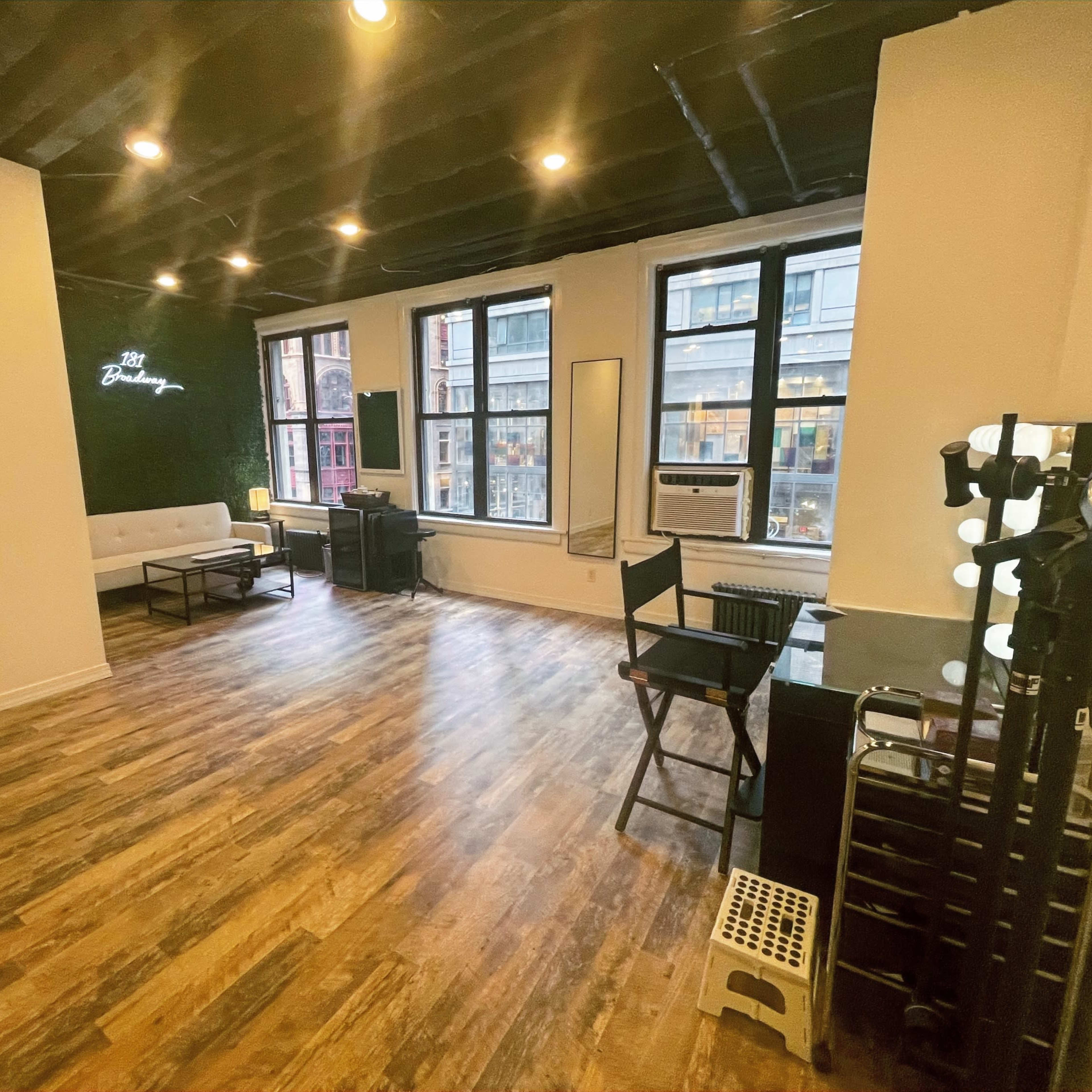 A spacious room featuring wooden flooring, large windows, and professional lighting equipment along with minimalist furniture.