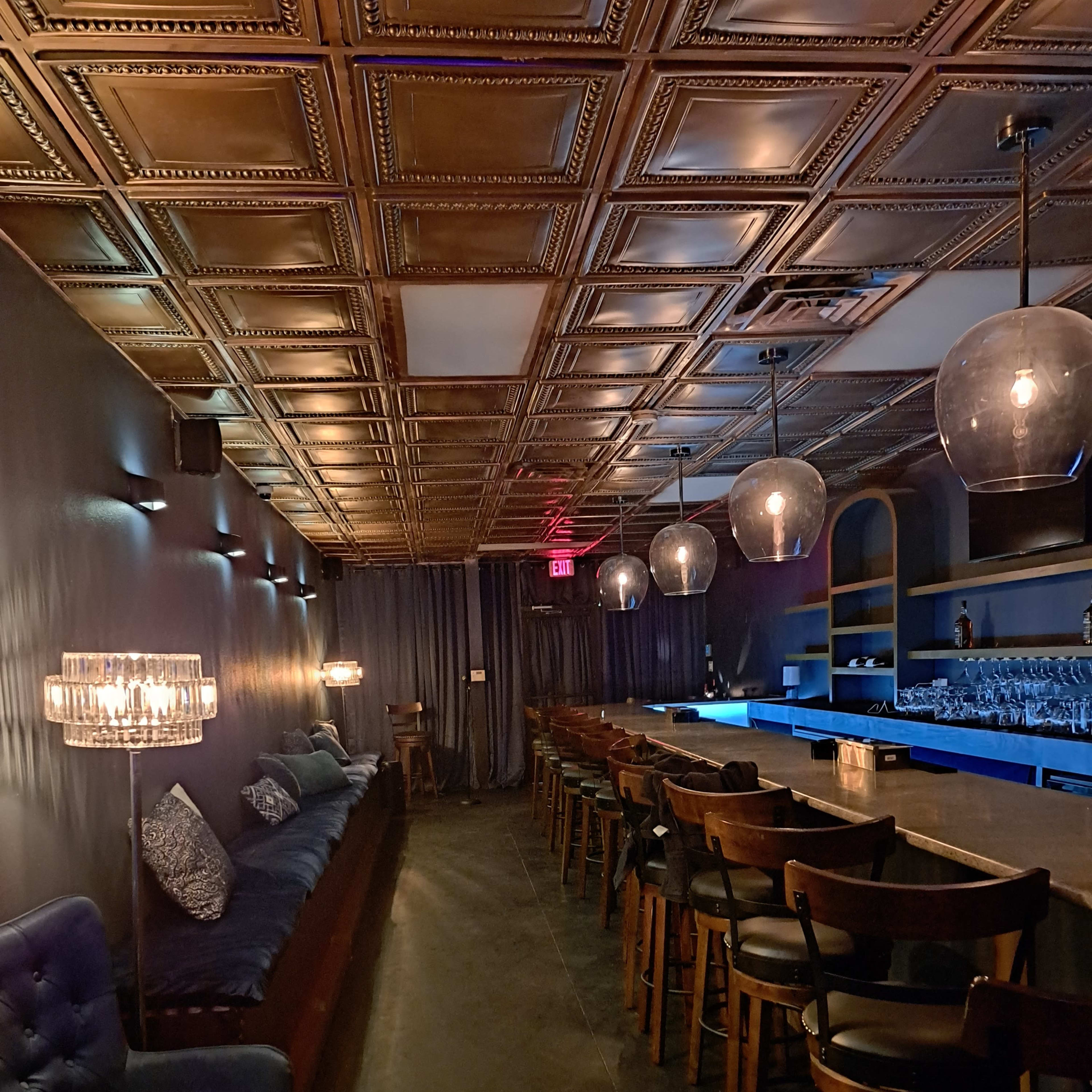 The interior of a dimly lit lounge features a long bar with wooden seating, plush chairs, and overhead pendant lights illuminating a stylish decor.