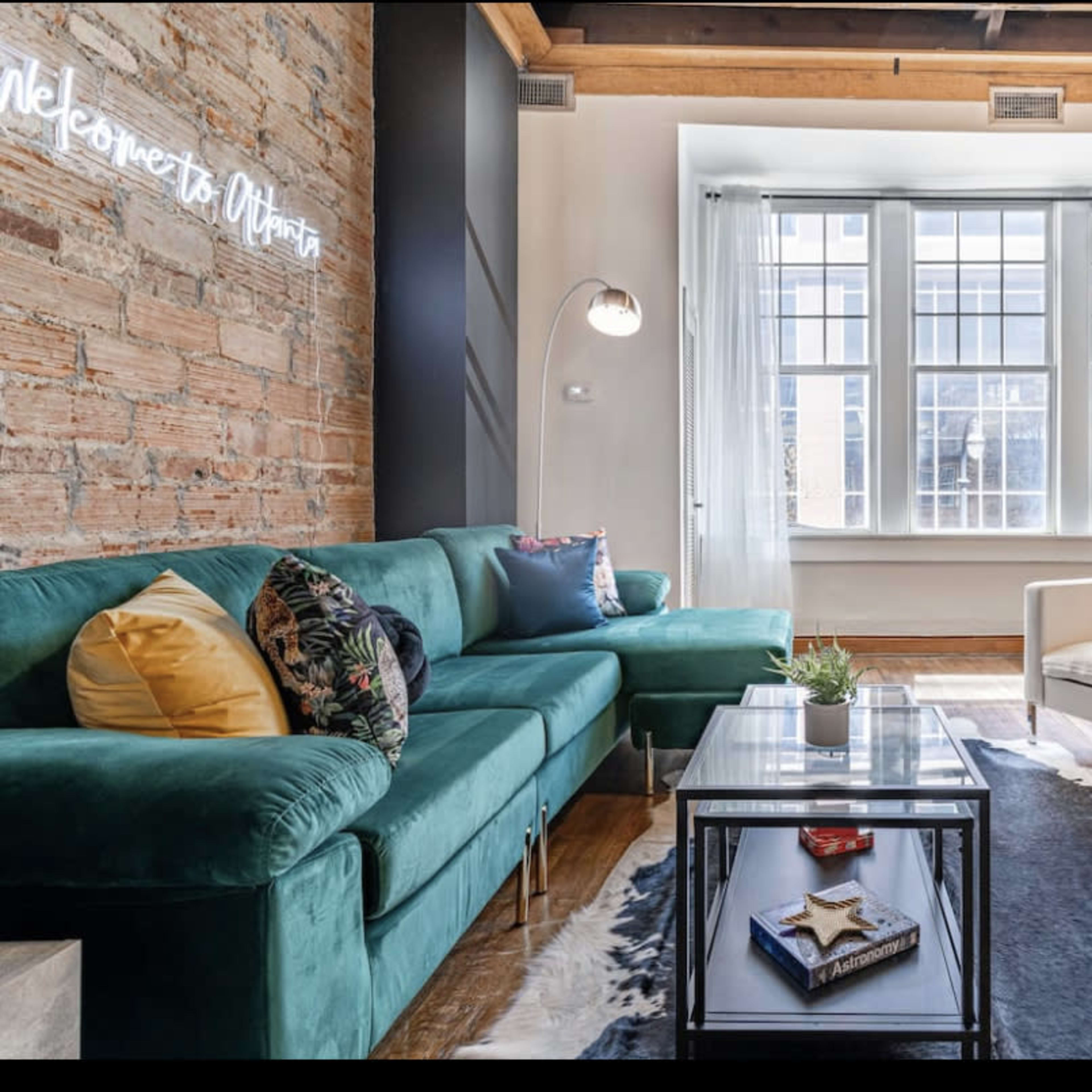 The living room features a green velvet sofa, a white armchair, a glass coffee table, and large windows allowing natural light to fill the space.