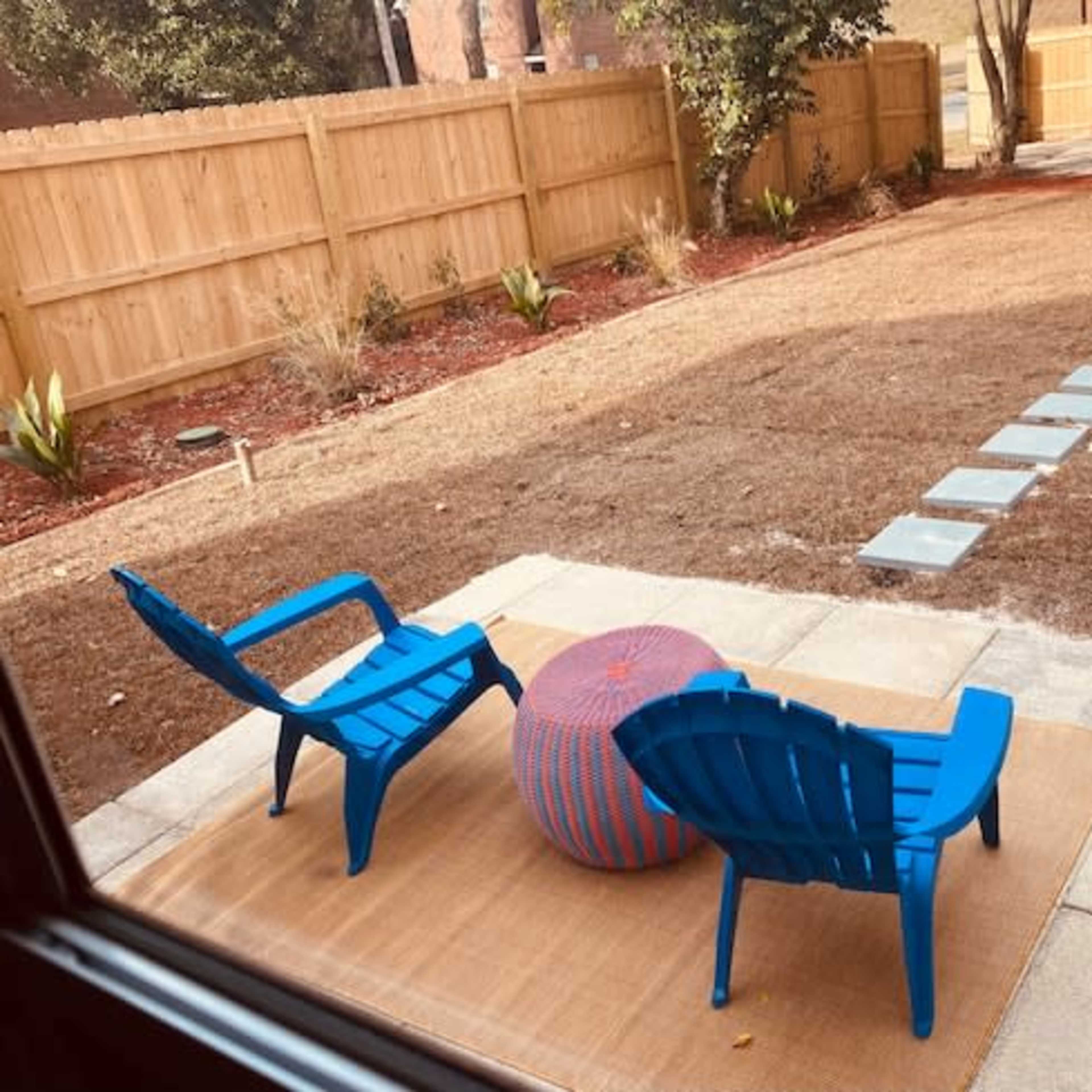 A backyard with two blue chairs, a circular patterned ottoman, and a pathway of stone slabs leading across a brown grassy area towards a fence.
