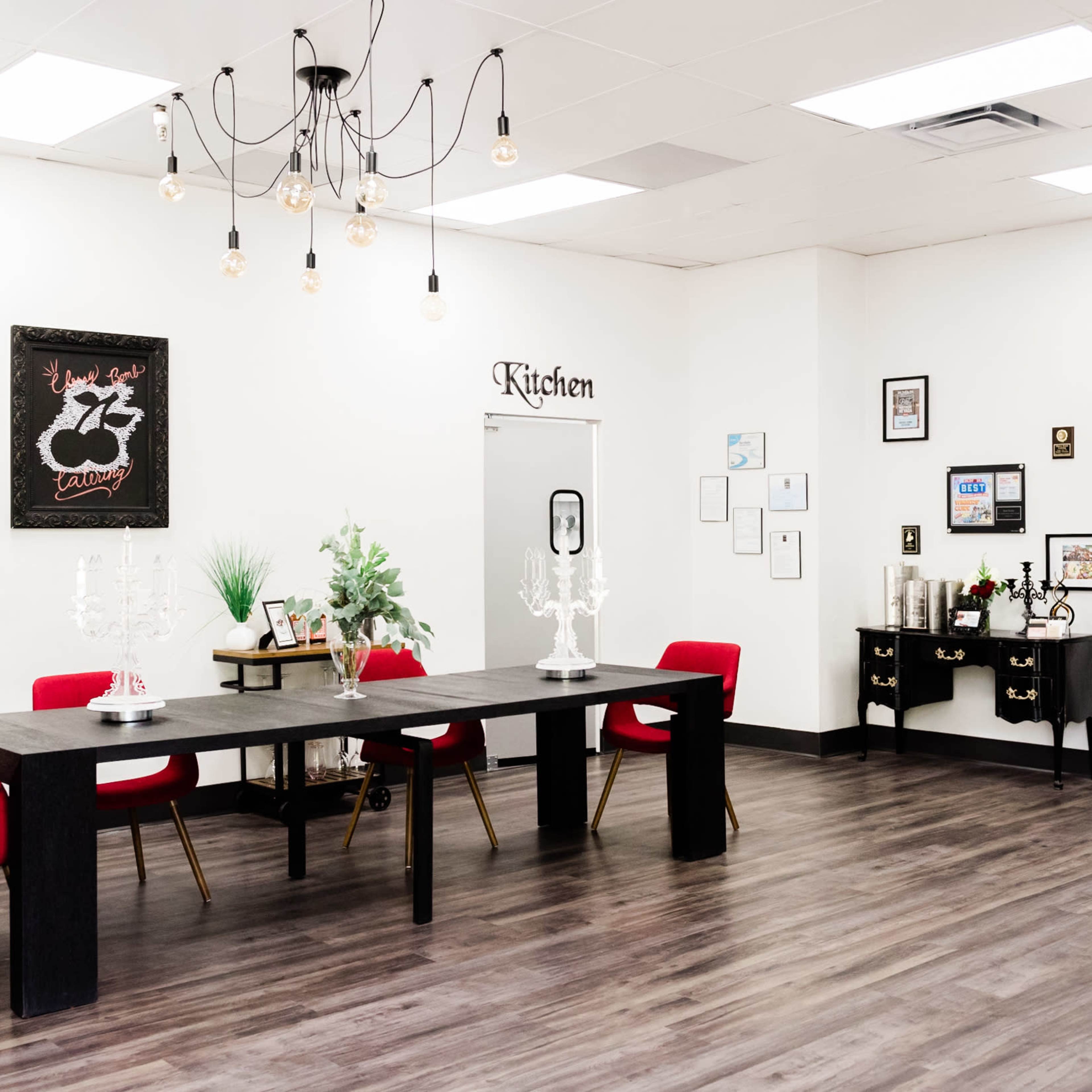 A modern, well-lit kitchen space featuring a long black table surrounded by red chairs, greenery, and decorative accents on the walls.