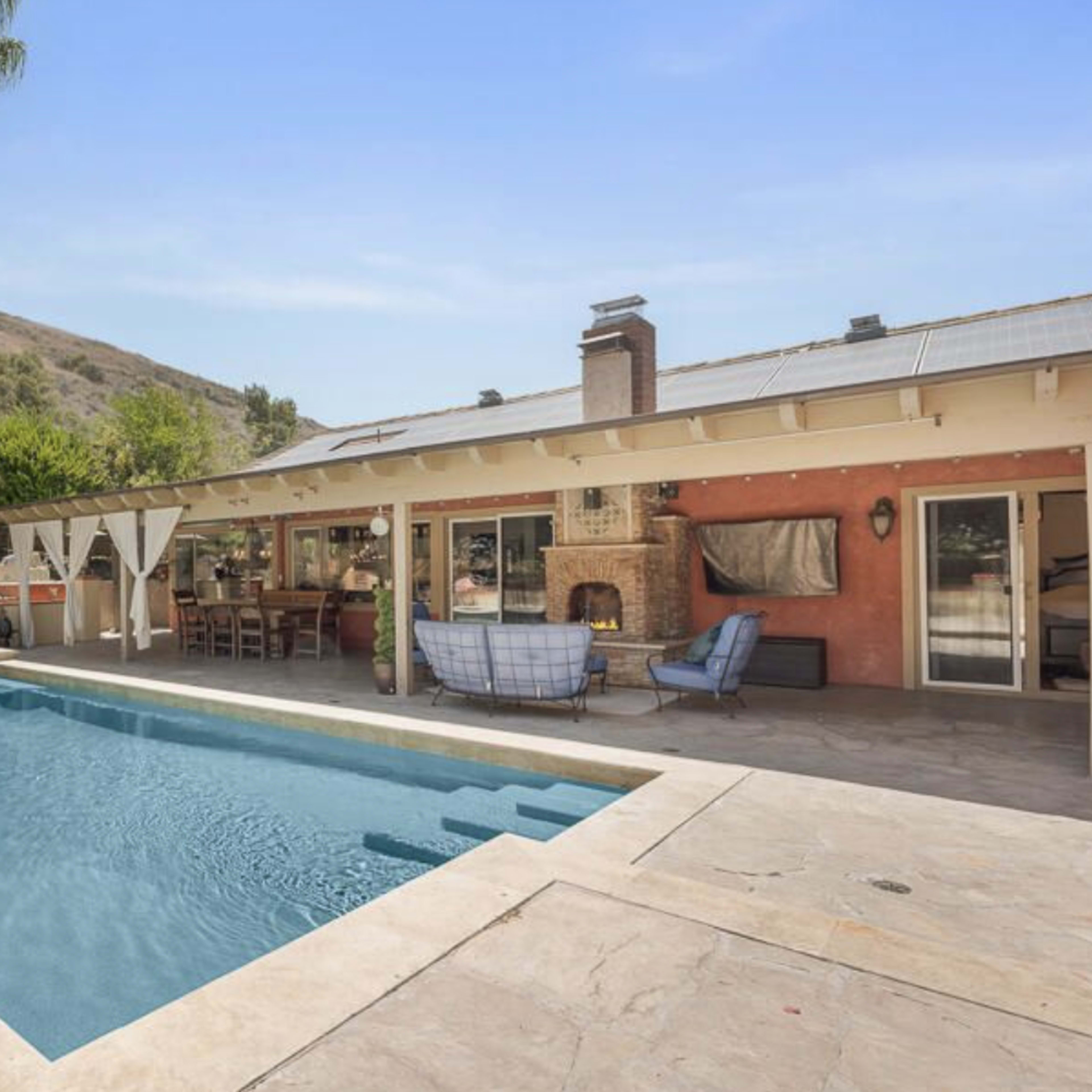 A pool area with a patio and outdoor seating adjacent to a house surrounded by trees and hills.