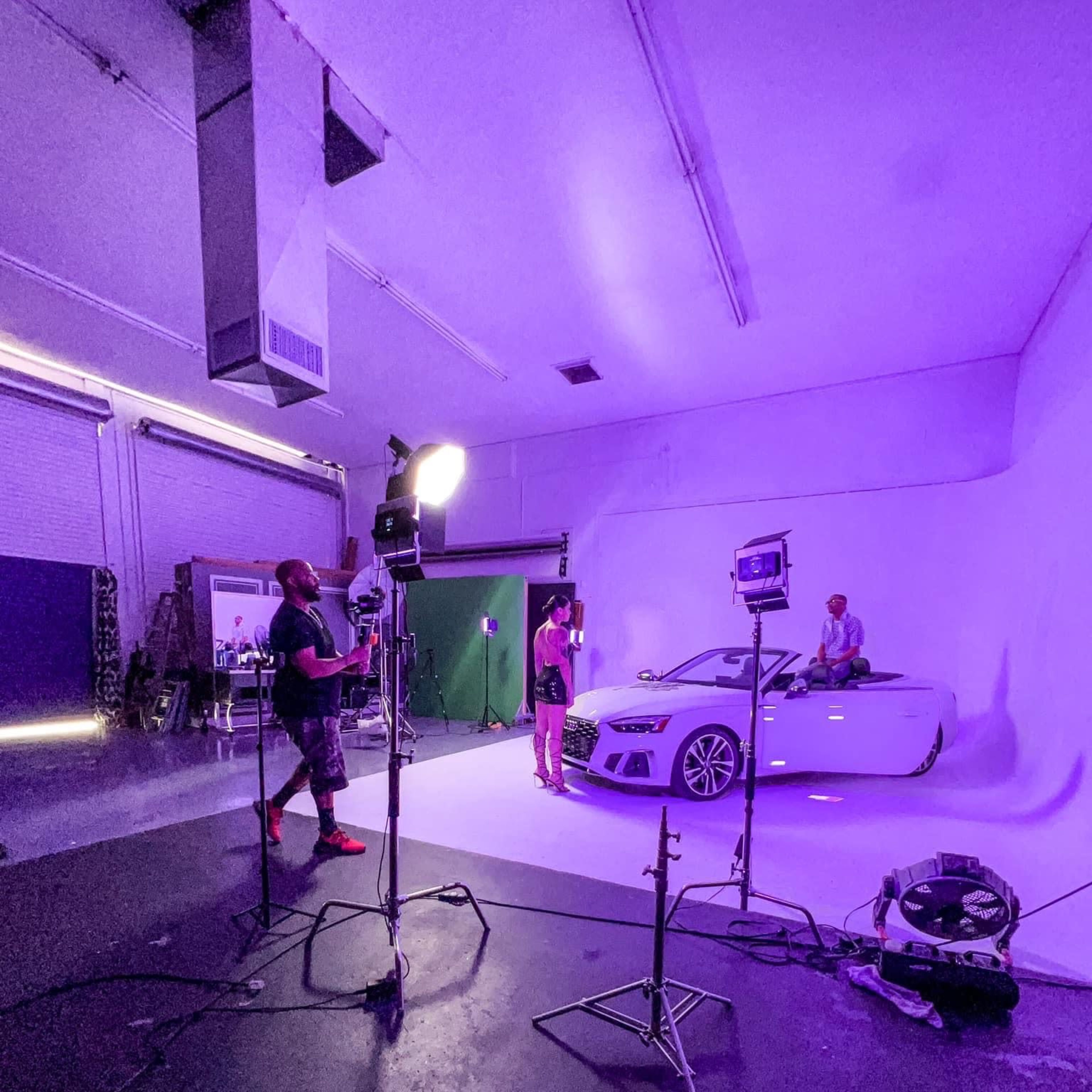 A photo shoot setup in a studio with a white car, lights, and crew members preparing for a shoot against a purple backdrop.