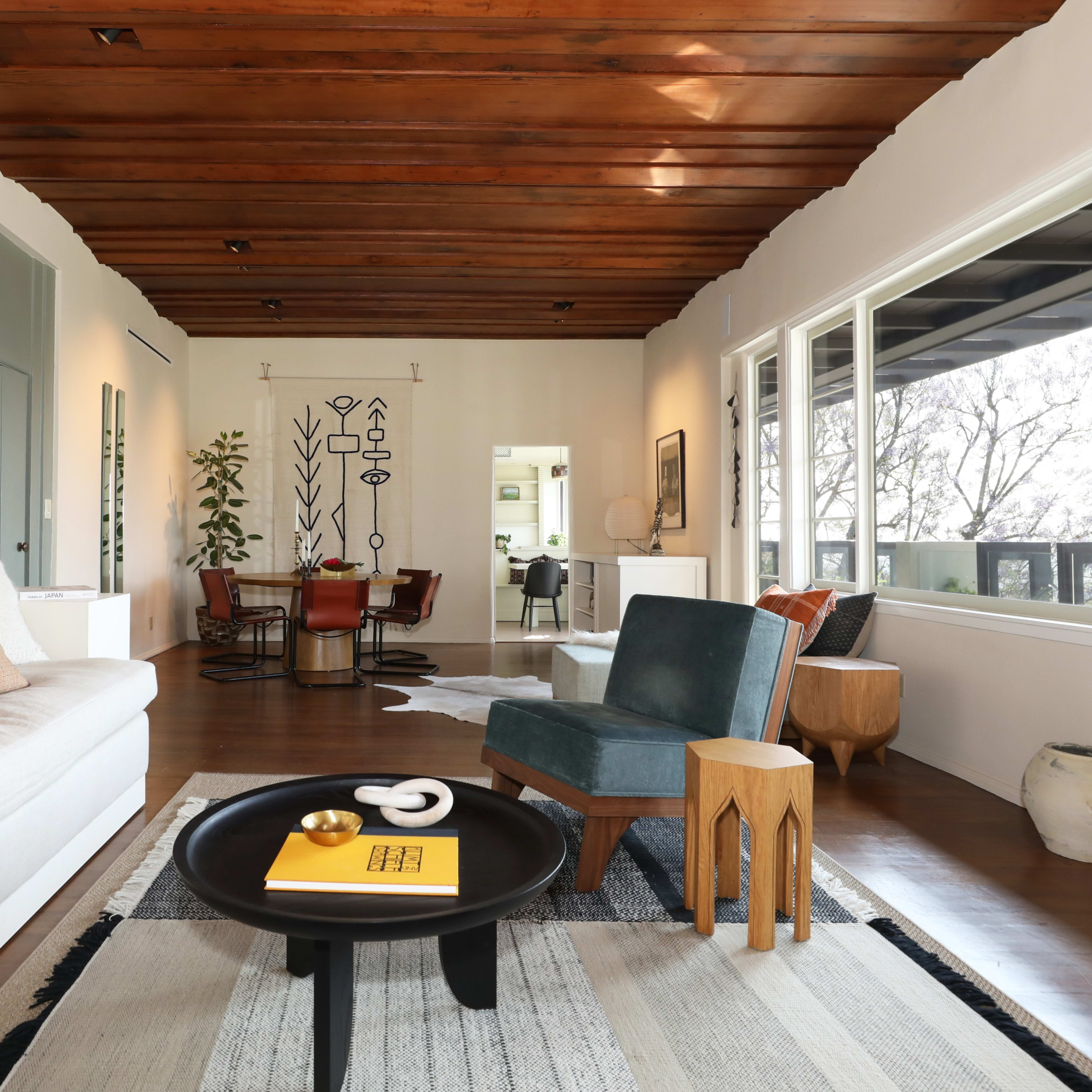 A modern living room with a light-colored sofa, wooden beam ceiling, and a coffee table, leading to an adjacent dining area with a large window.