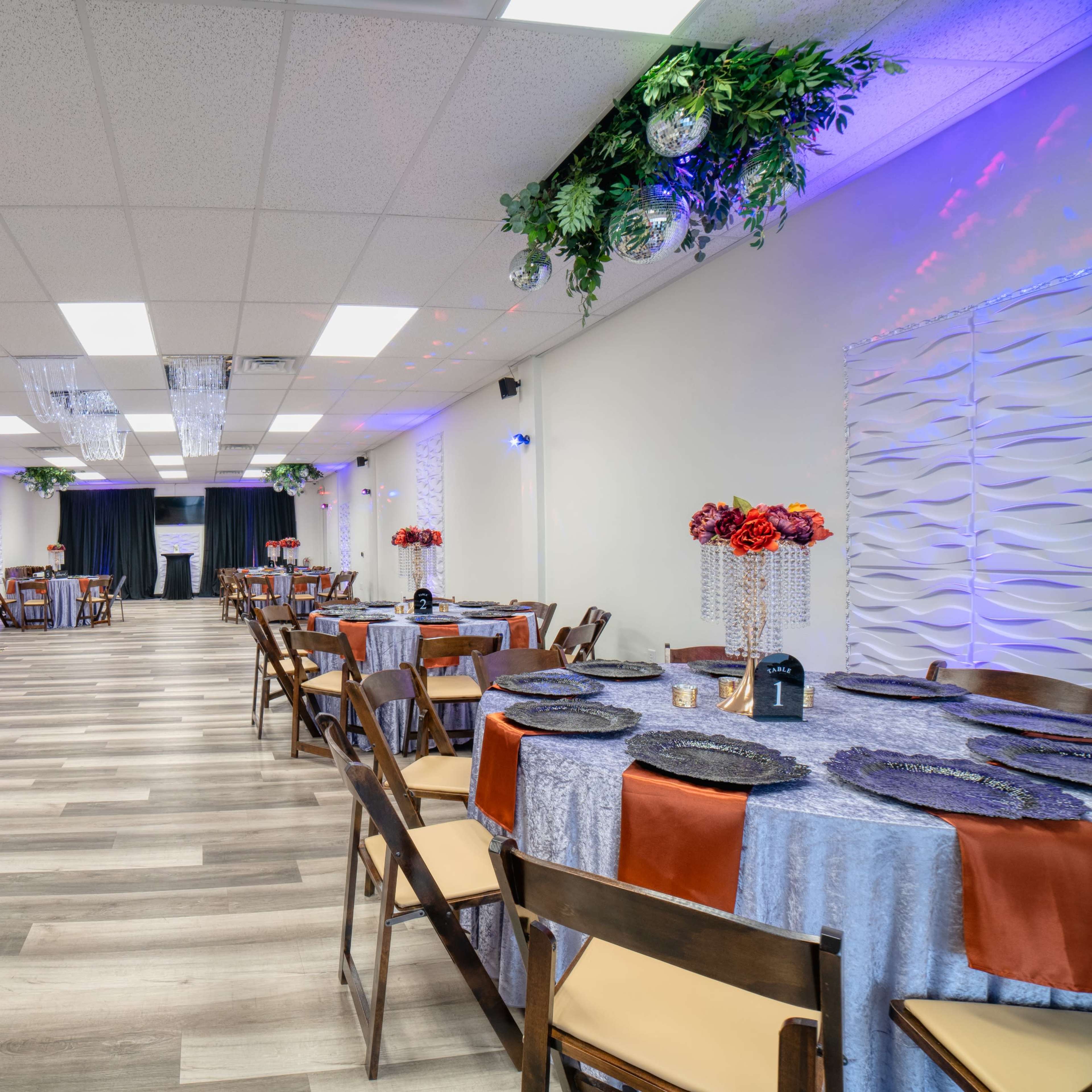 A spacious event room features tables with decorative coverings, arranged with chairs, and accented by floral centerpieces and ambient lighting.