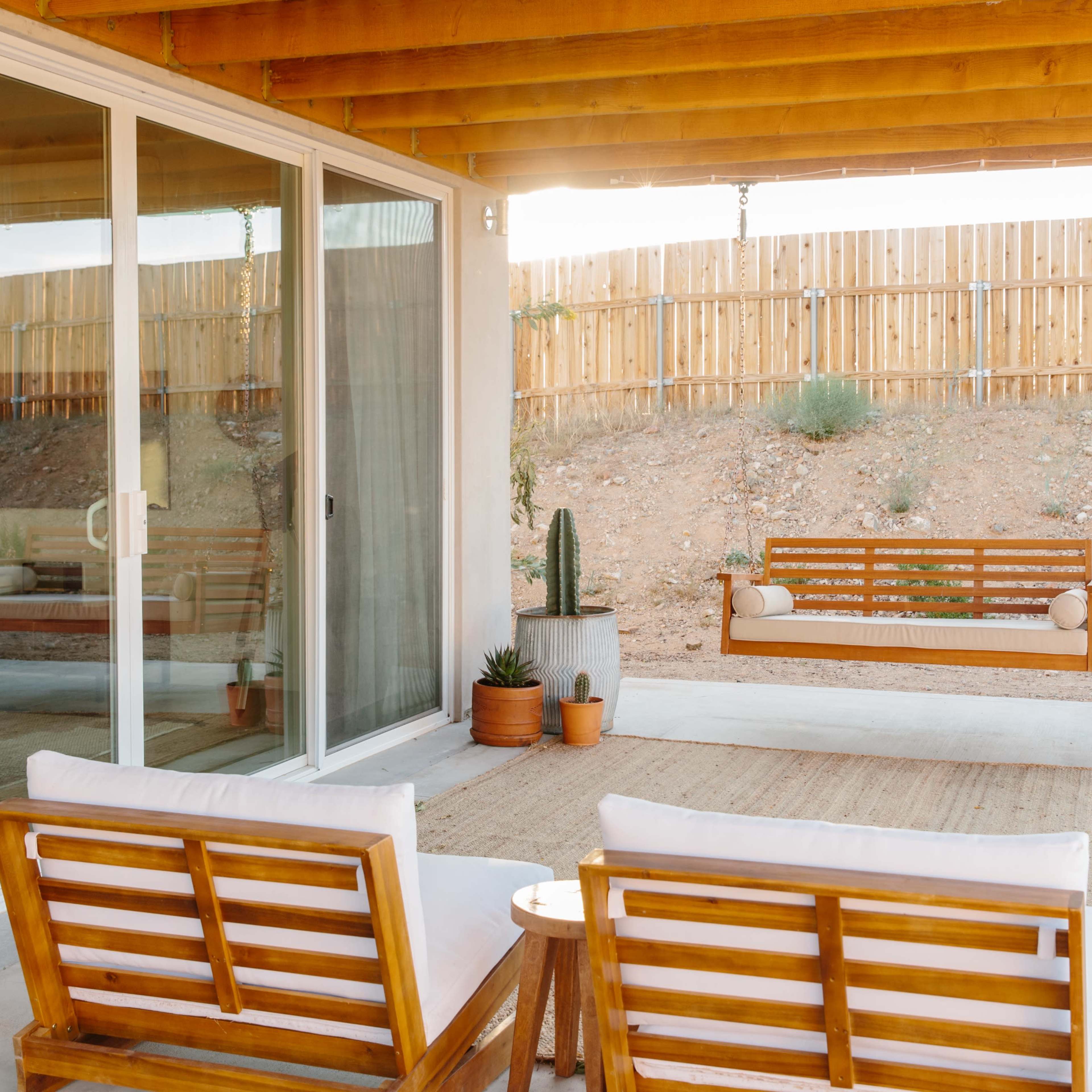 A patio area features two wooden chairs and a swing, with potted plants and a wooden fence in the background.