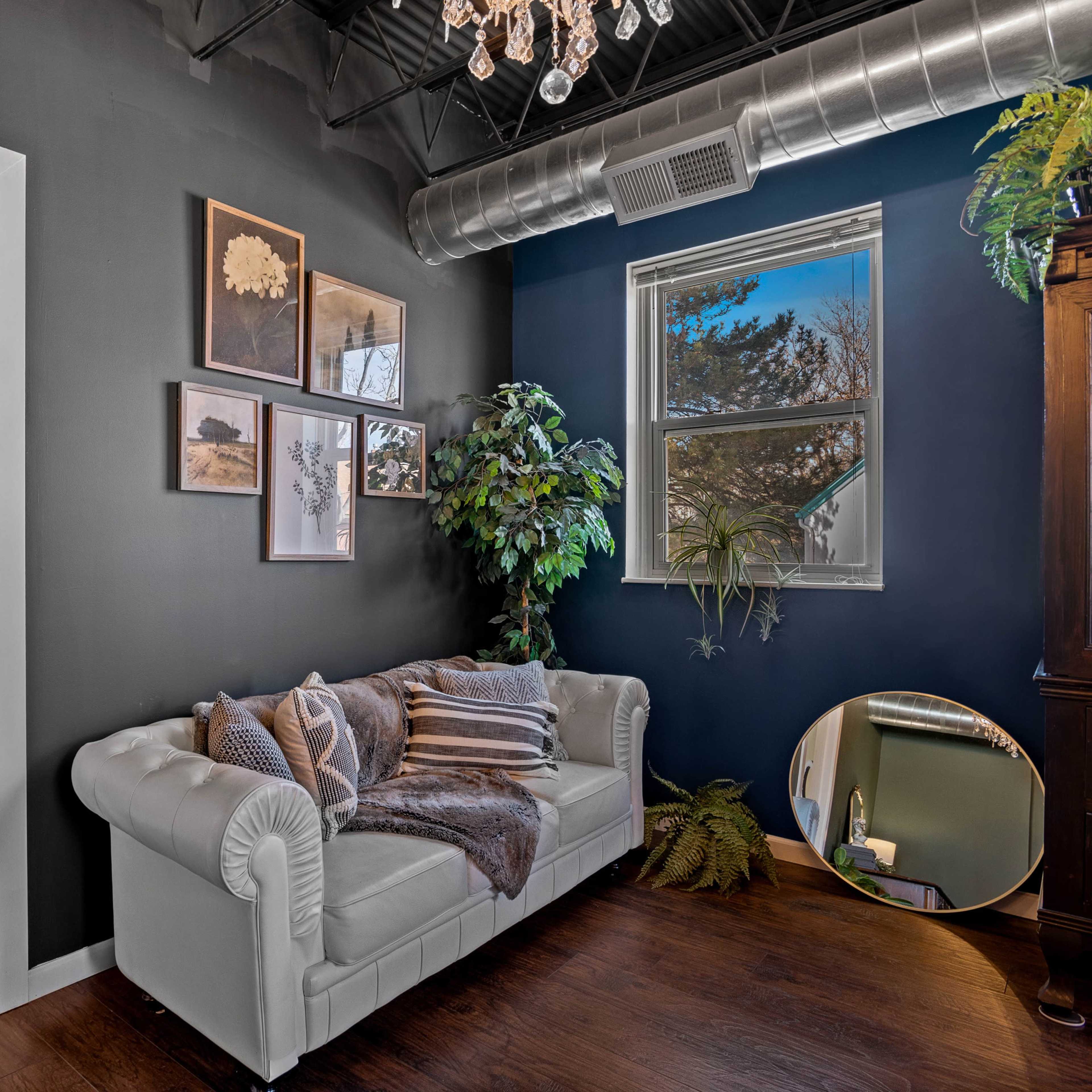 A modern living space with a white couch, dark blue walls, framed pictures, and a wall-mounted mirror, alongside a doorway leading to another room.