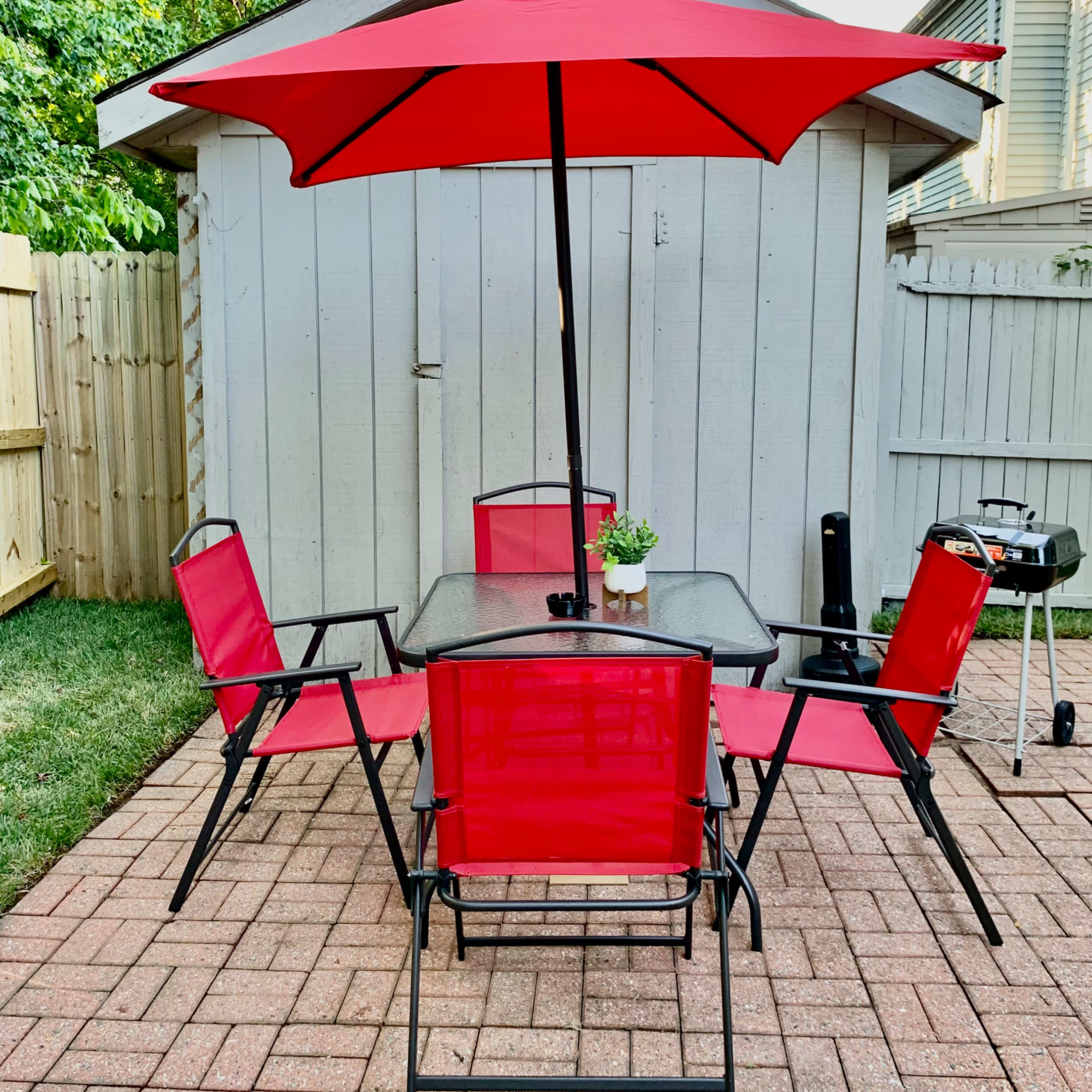 A patio area features a glass table surrounded by four red chairs, topped with a red umbrella, adjacent to a wooden shed.