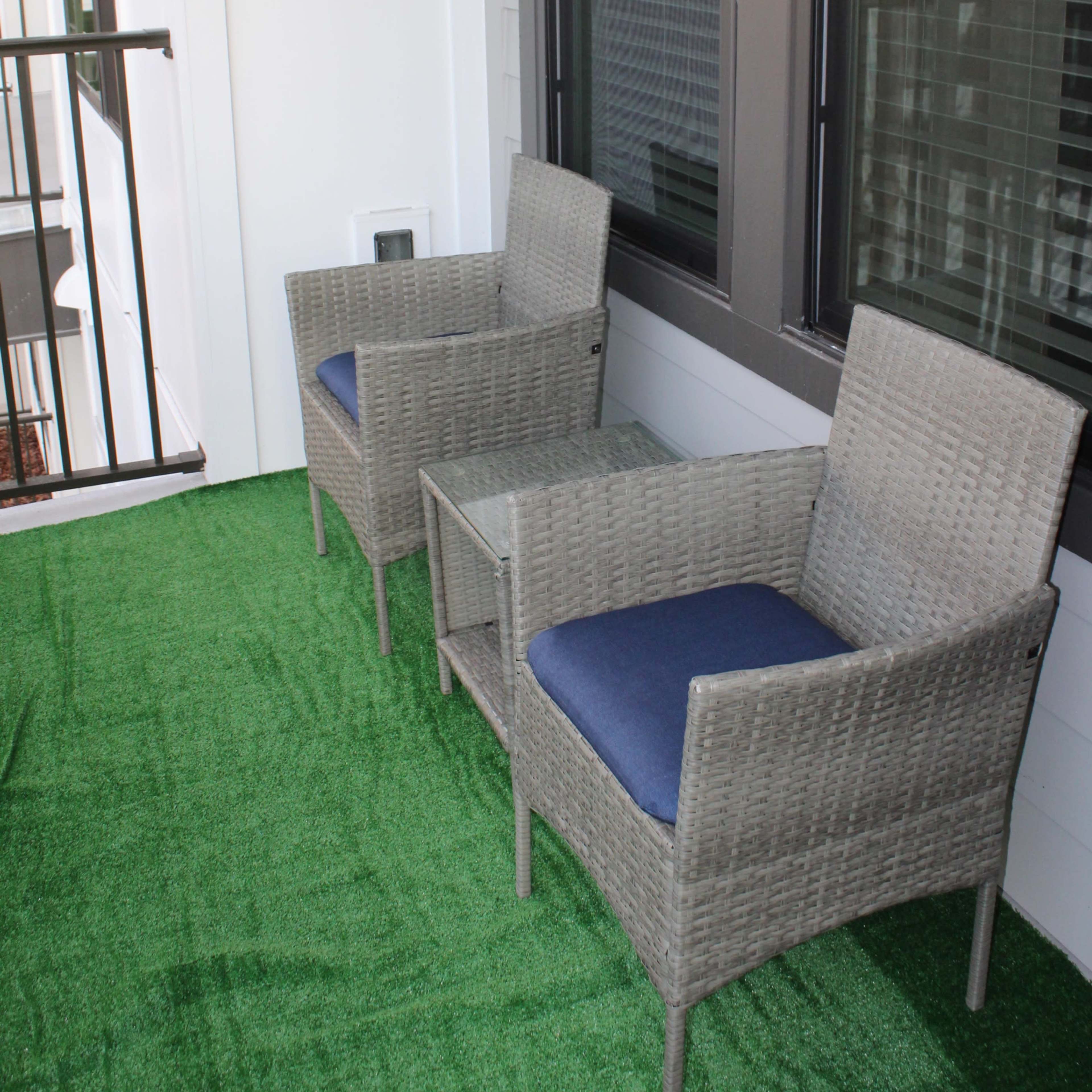 A small balcony with two wicker chairs and a table, surrounded by green synthetic grass.