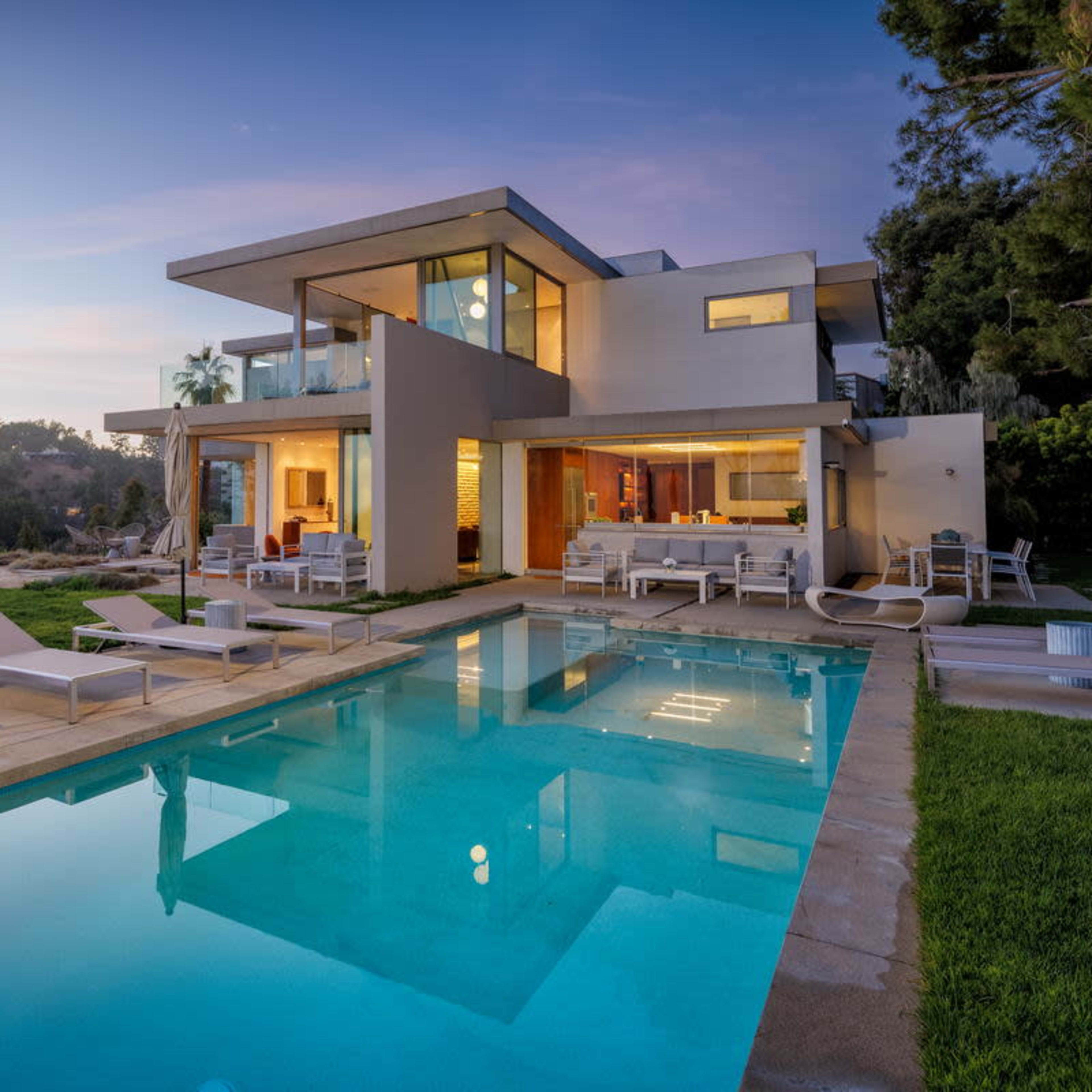 A modern two-story villa with large glass windows overlooks a swimming pool surrounded by lounge chairs and shaded by umbrellas, set against a twilight sky.