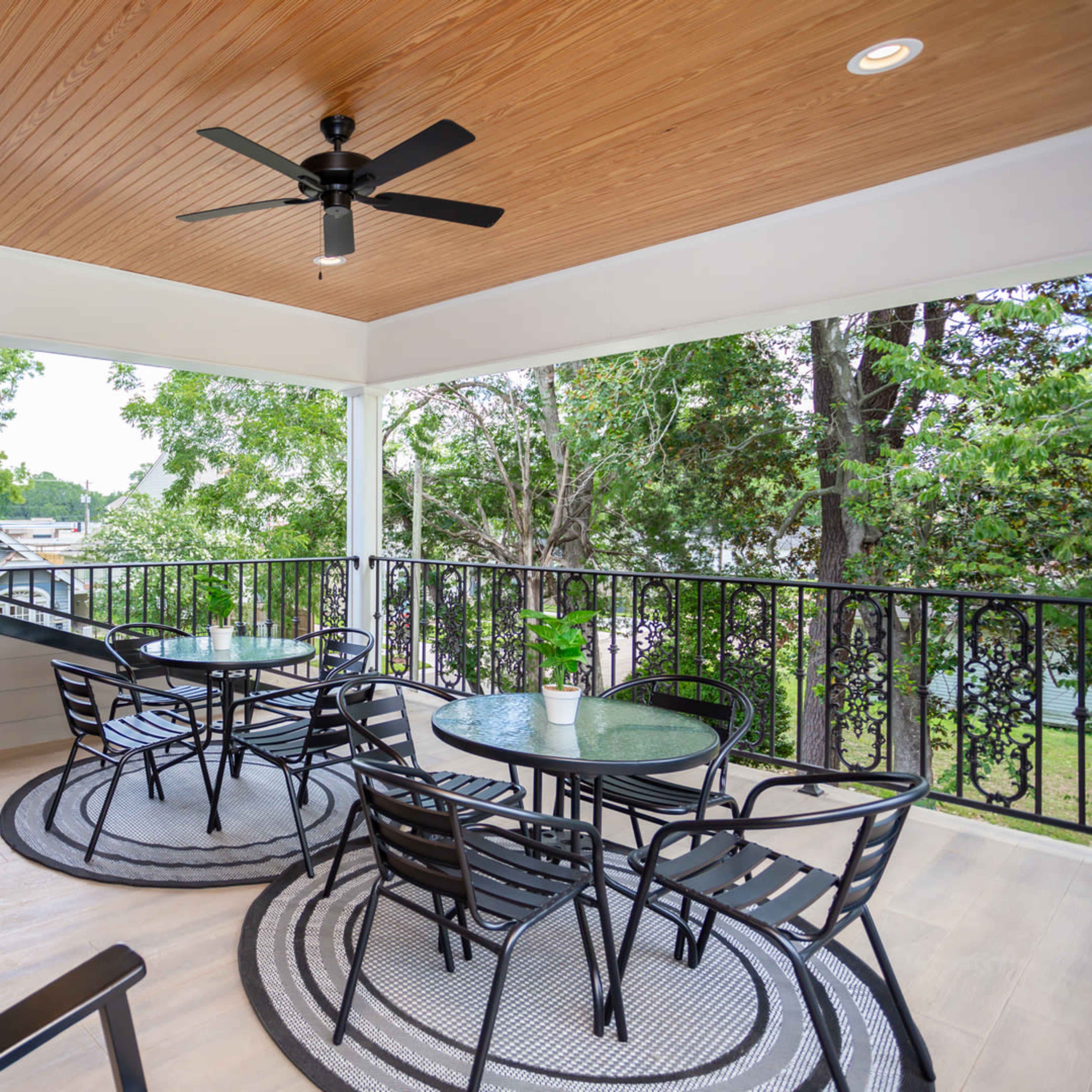 A covered patio features black metal furniture and a ceiling fan, surrounded by trees and a railing.