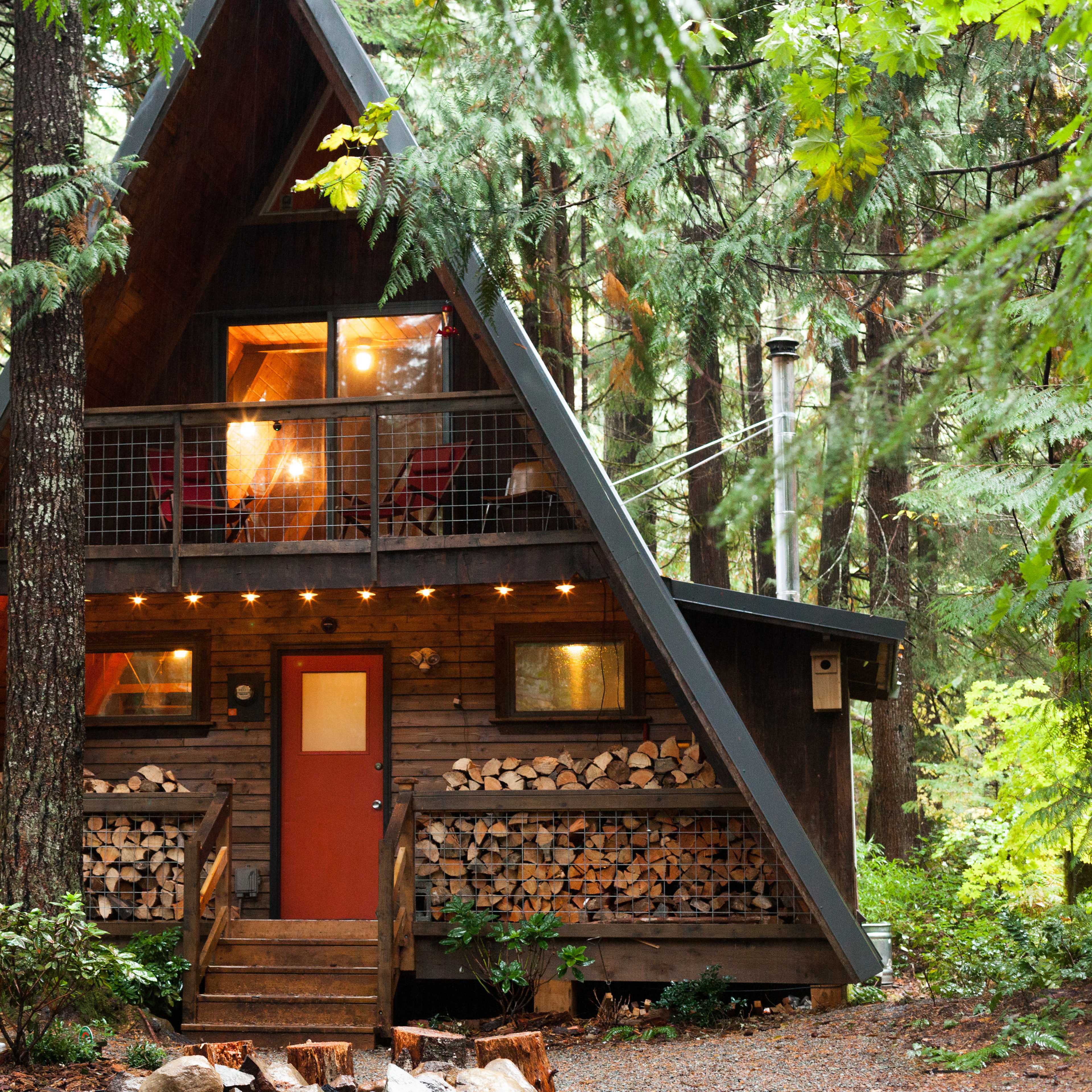 A wooden A-frame cabin with a red door is surrounded by tall trees and features stacked firewood on its front porch.