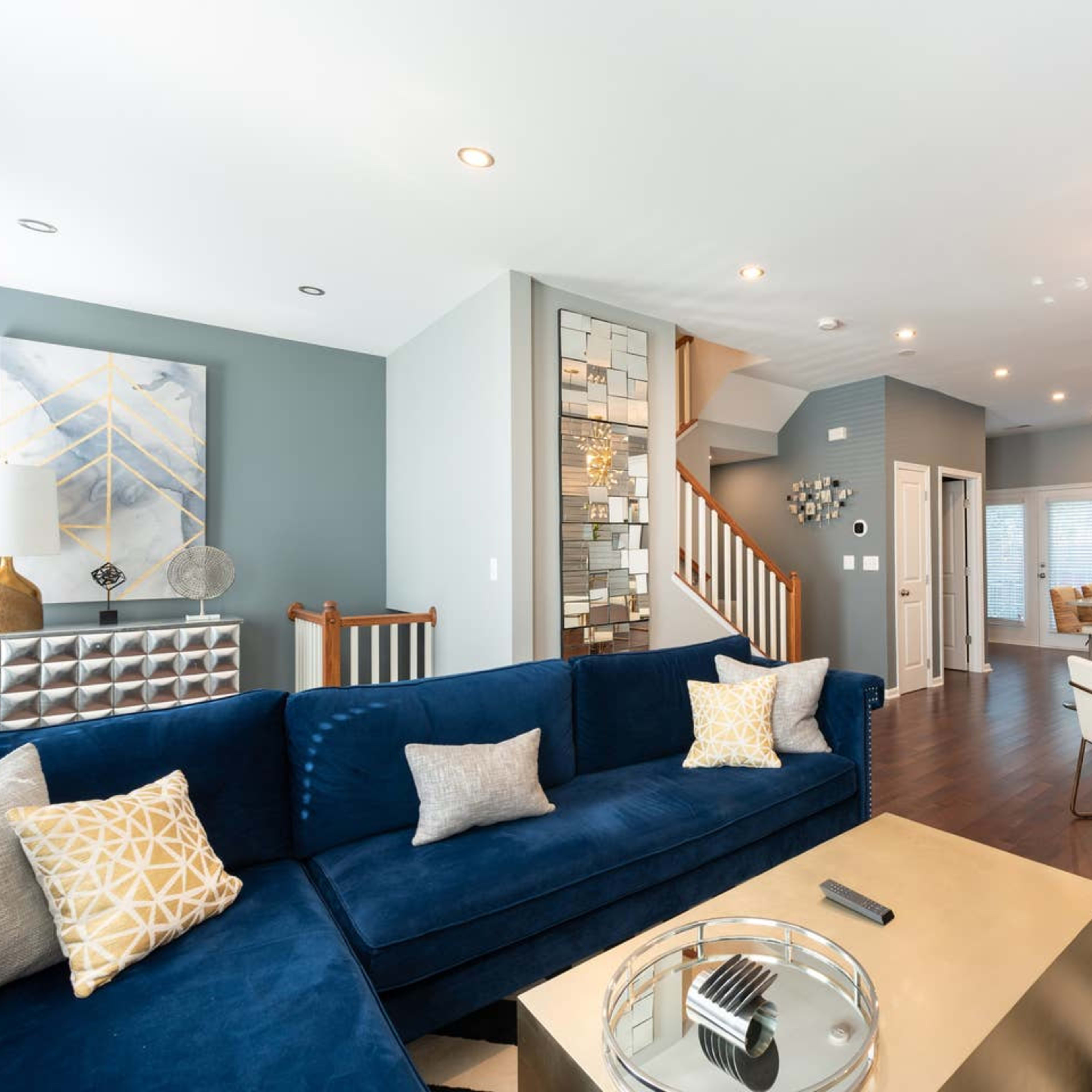 A modern living room featuring a blue velvet sofa, a gold coffee table, and a dining area with white chairs, illuminated by a stylish light fixture.