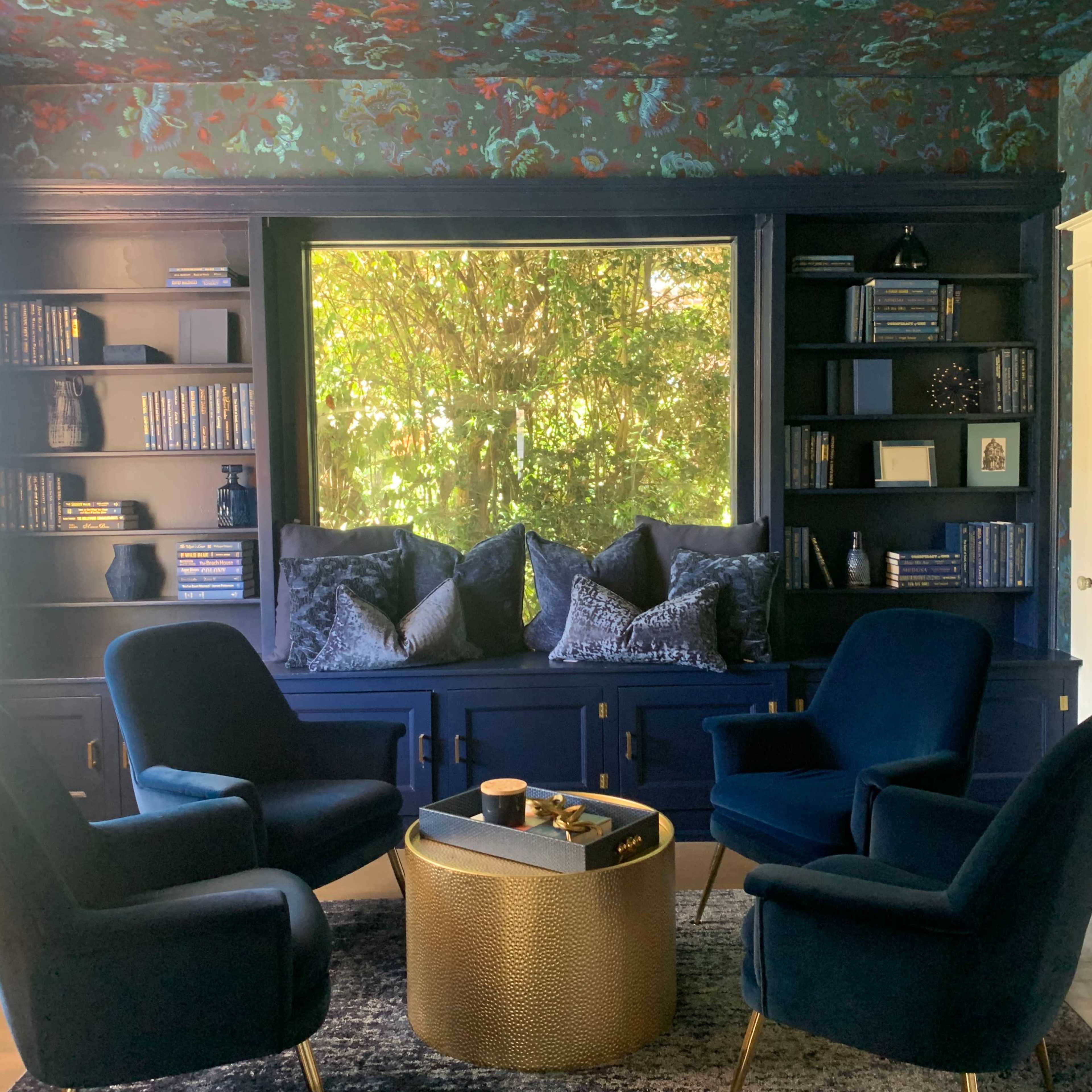 A cozy seating area features four blue chairs arranged around a metallic gold coffee table, with bookshelves and a large window adorned with leafy views in the background.