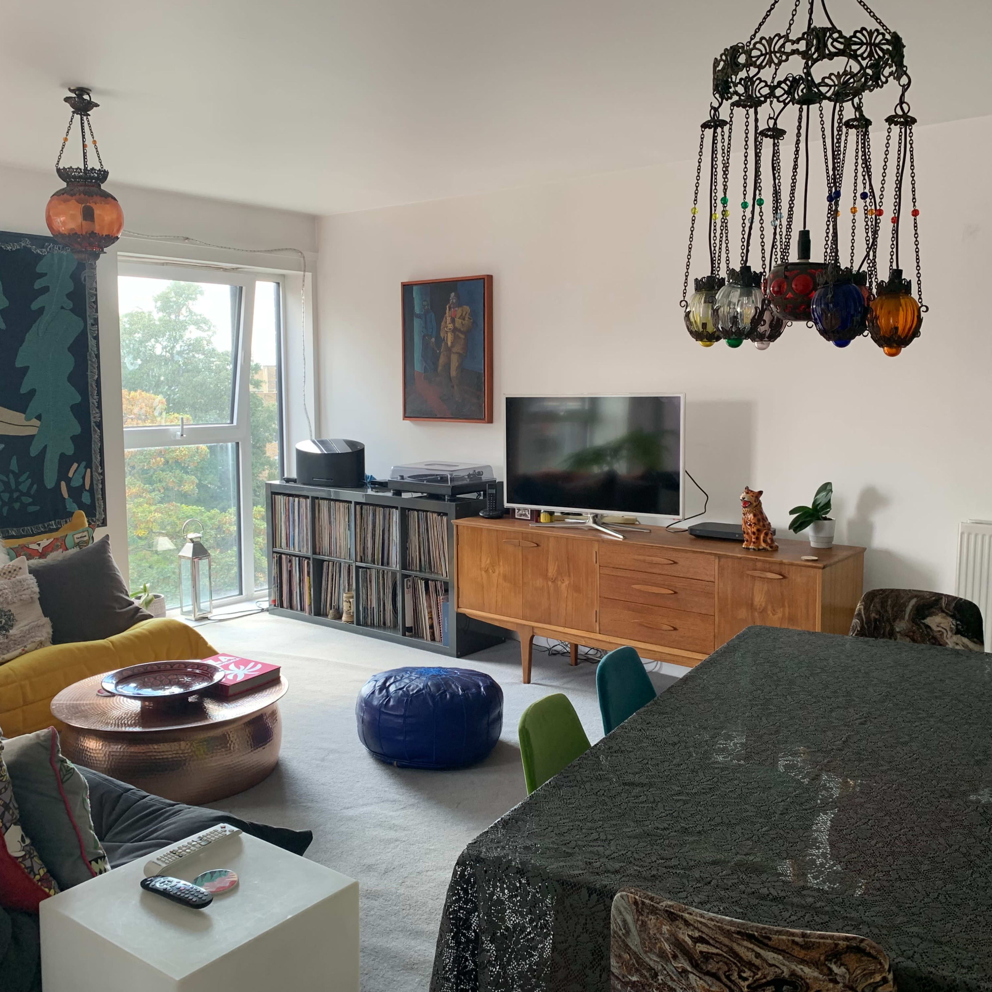 A living room features a large dining table, colorful chairs, a television on a wooden cabinet, and decorative lighting hanging from the ceiling.